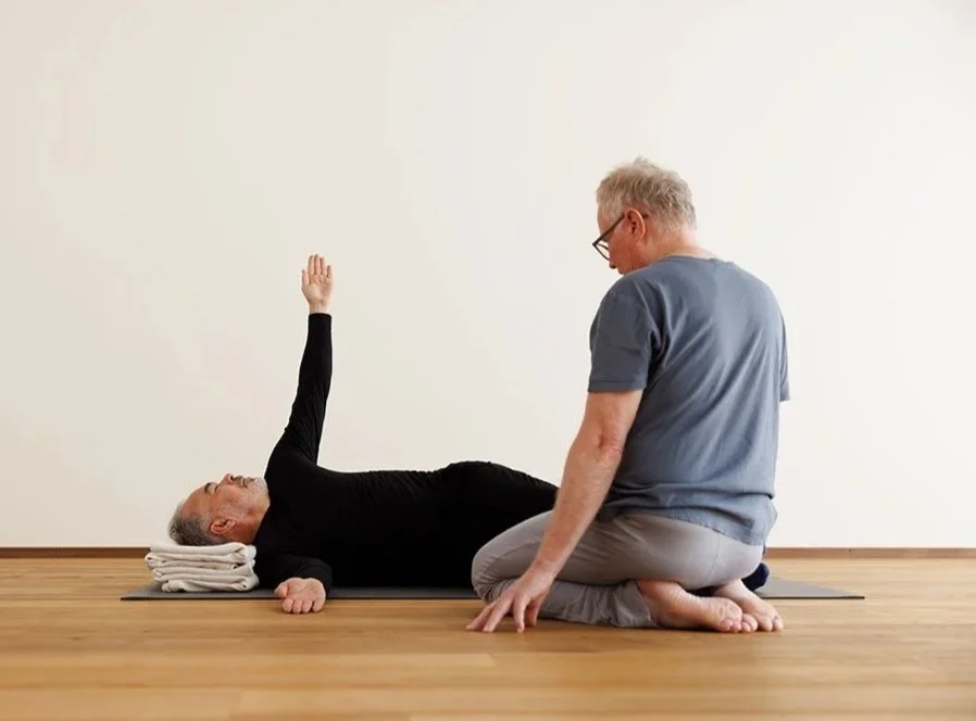 Zwei ältere Männer, einer liegt auf einem Yoga-Matte mit Kopf auf gerollten Handtüchern, der andere sitzt im Schneidersitz und führt eine Yogapose aus.