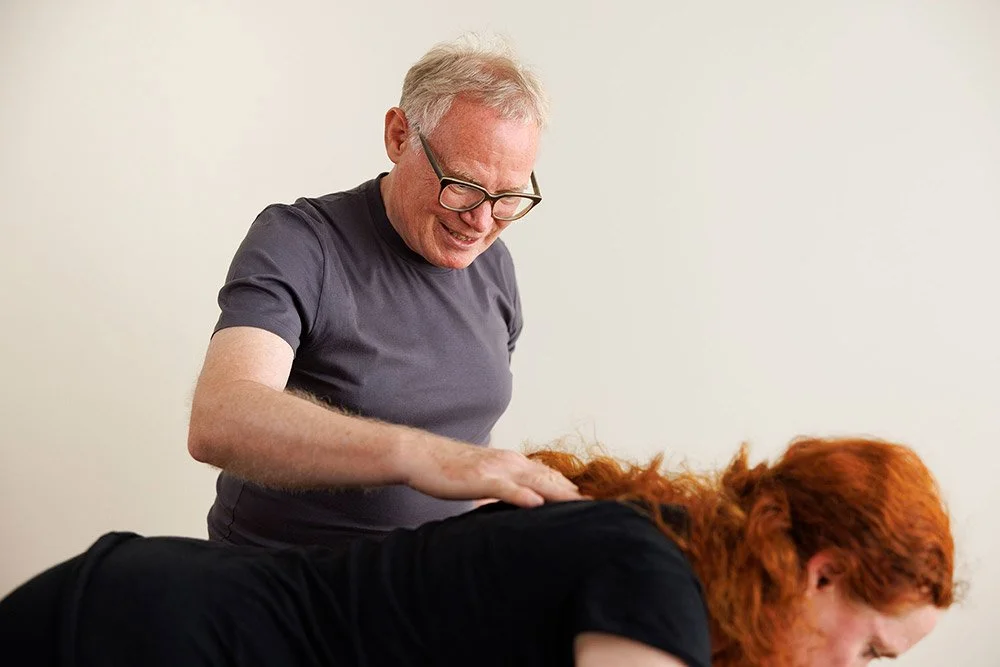 Ein Mann mit Brille, der eine Frau mit roten Haaren bei einer Yoga-Übung unterstützt.