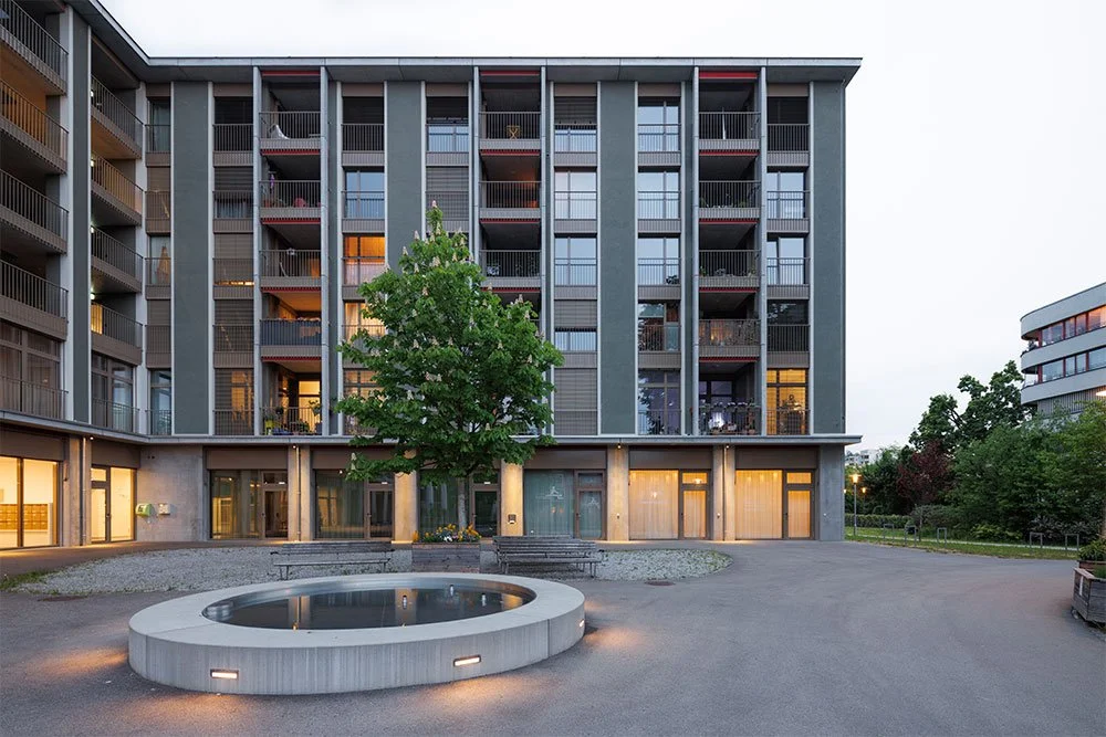 Mehrstöckiges modernes Wohngebäude mit Balkonen, in der Mitte ein Baum, im Vordergrund ein rundes Wasserbecken, umgeben von Wegen und Bänken, bei Dämmerung.