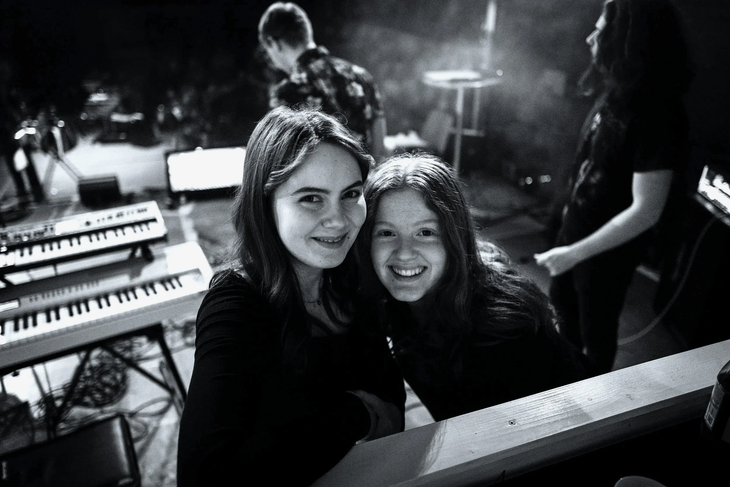 Zwei lachende junge Frauen, die sich für ein Selfie in einem Musikstudio befinden, mit Keyboards und anderen Musikern im Hintergrund.
