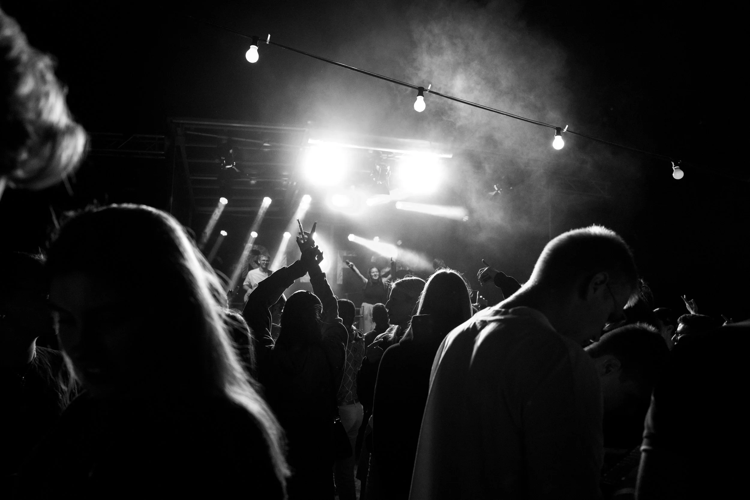 Bühne bei einem Konzert in Schwarz-Weiß, mit mehreren Menschen im Publikum, einige mit erhobenen Händen, auf der Bühne sind Sänger und Musiker mit Licht- und Scheinwerfereffekten.