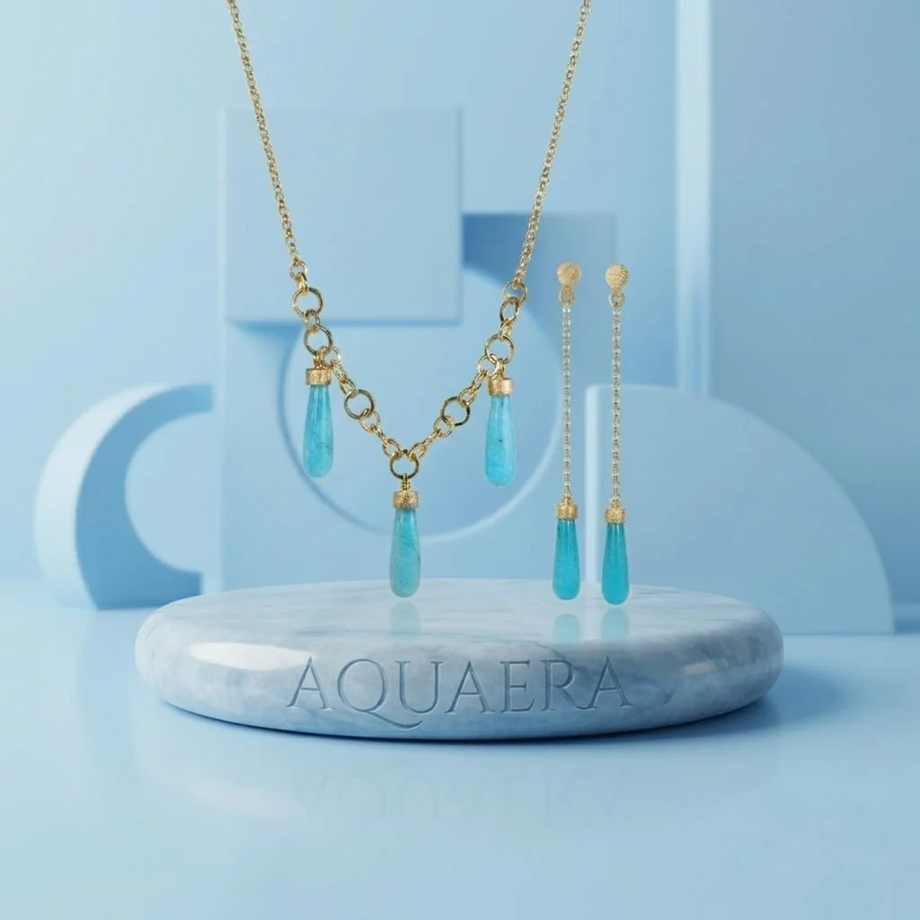 Gold-plated necklace and earrings with blue agate teardrop pendants, displayed on a marble stand with the brand name "AQUAERA" against a blue geometric background.
