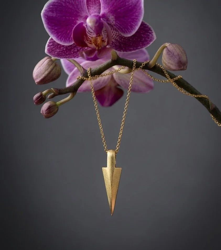 A gold-plated necklace with a spike-like pendant hanging from a pink orchid flower with buds, against a dark gray background.