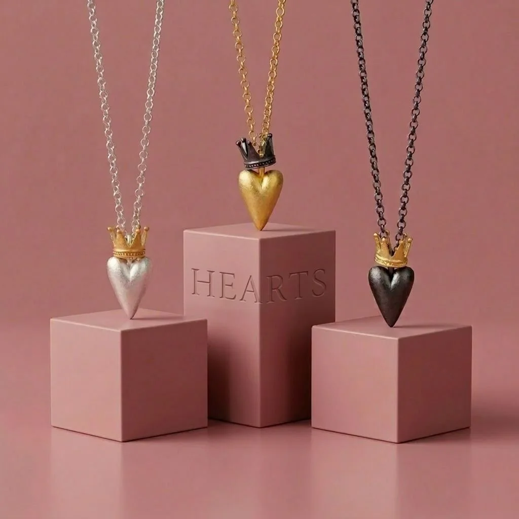 Three necklaces with heart-shaped sterling silver, gold-plated, ruthenium-plated pendants, each wearing a small crown, displayed on pink boxes against a pink background.