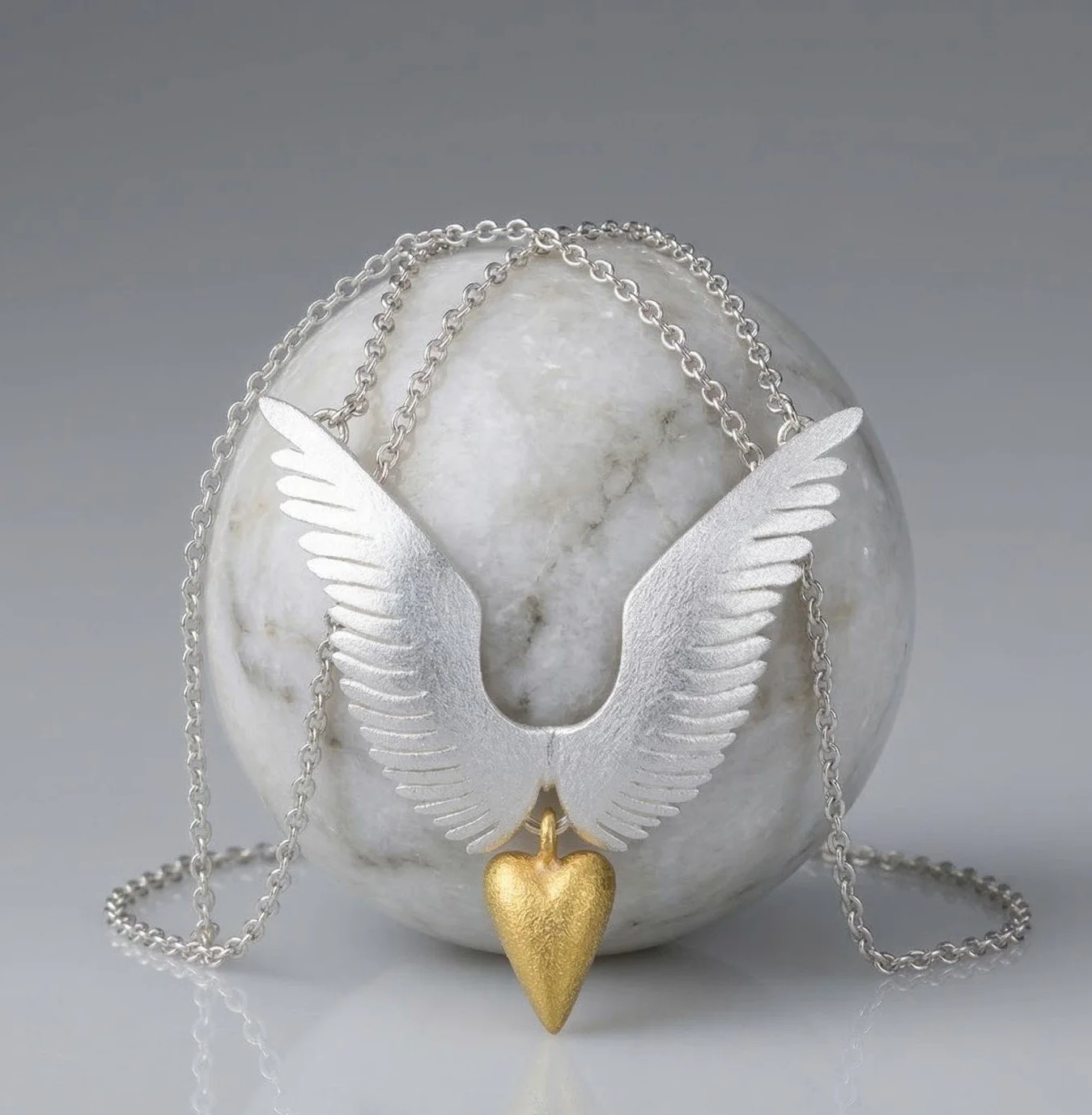 Sterling silver necklace with angel wings and a gold-plated heart pendant, draped over a white spherical object with a reflective surface.