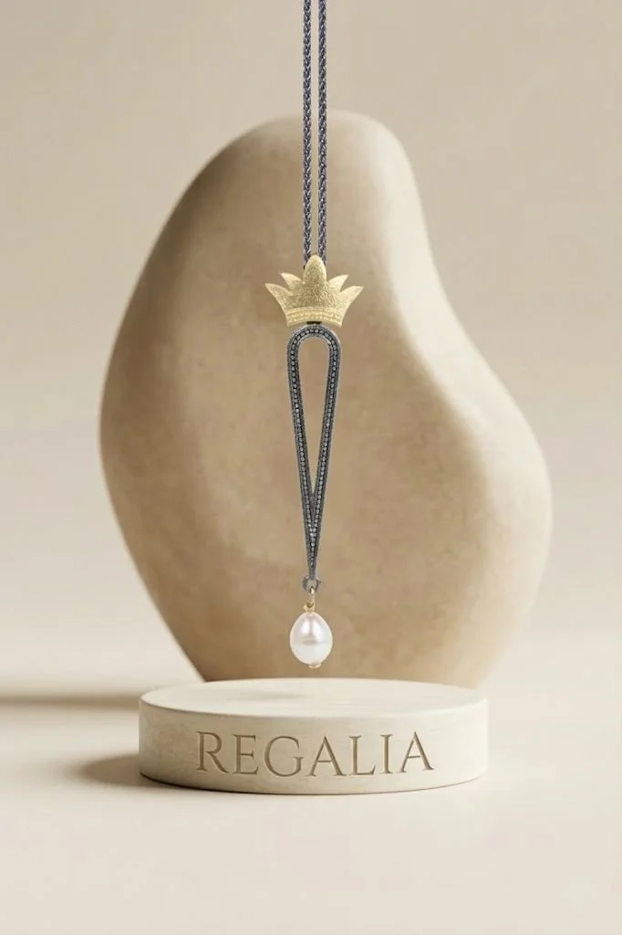 A ruthenium-plated pearl pendant necklace hanging from a crown-shaped gold-plated ornament, displayed in front of a smooth, beige stone with the word 'REGALIA' engraved on a circular base.