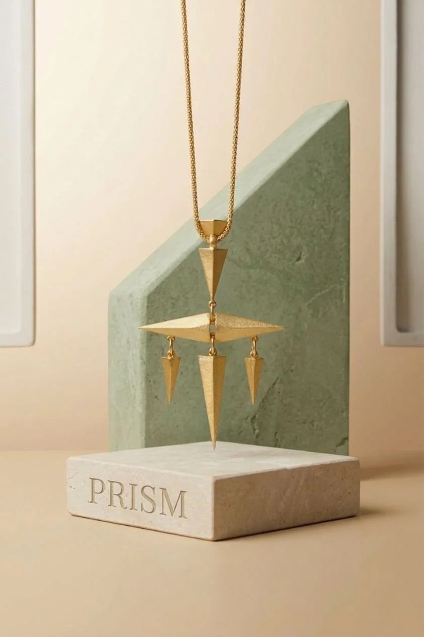 A gold-plated geometric pendant necklace with multiple spike-shaped pendants. The necklace hangs above a white marble block with the word "PRISM" engraved, with a green textured slab behind it.