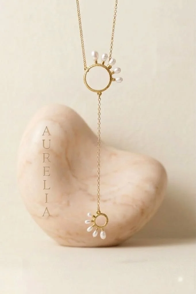 Gold-plated necklace with two circular pendants decorated with white pearls, hanging in front of a large beige shell with the name 'AURELIA' written vertically on it.