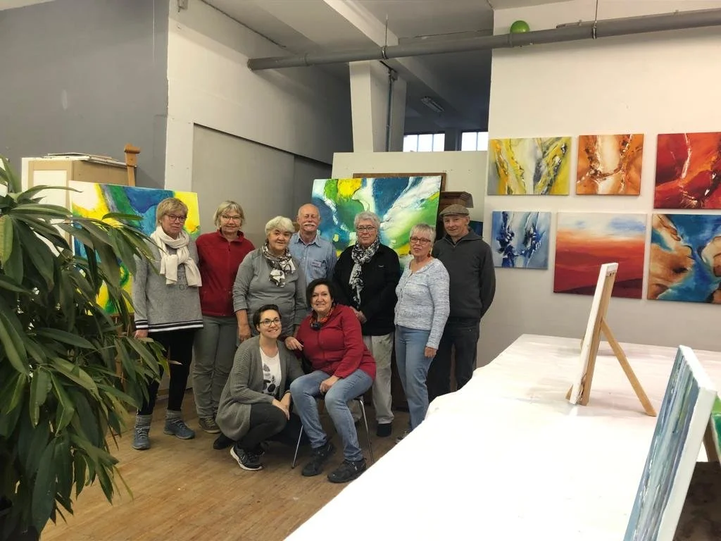 Eine Gruppe von Menschen steht in einer Kunstgalerie vor mehreren bunten abstrakten Gemälden an der Wand.