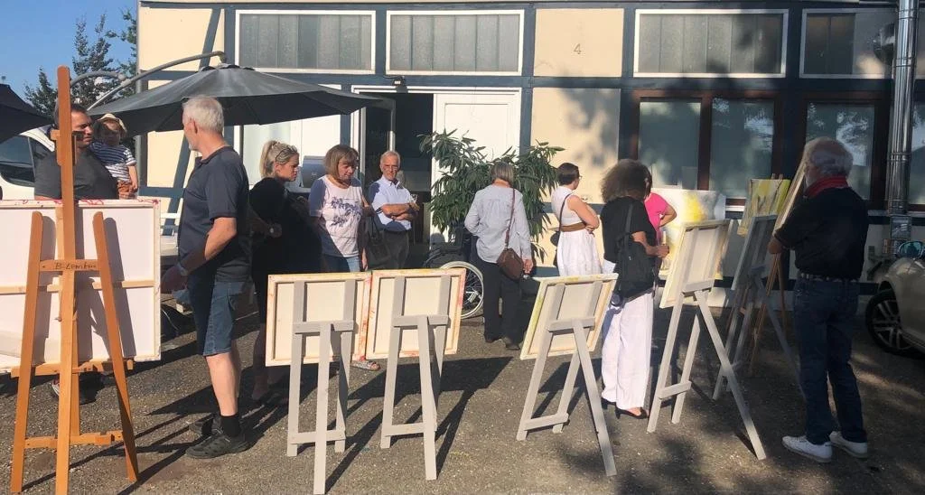 Mehrere Menschen stehen im Freien vor einem Gebäude und betrachten Bilder auf Staffeleien bei einem Kunstveranstaltung