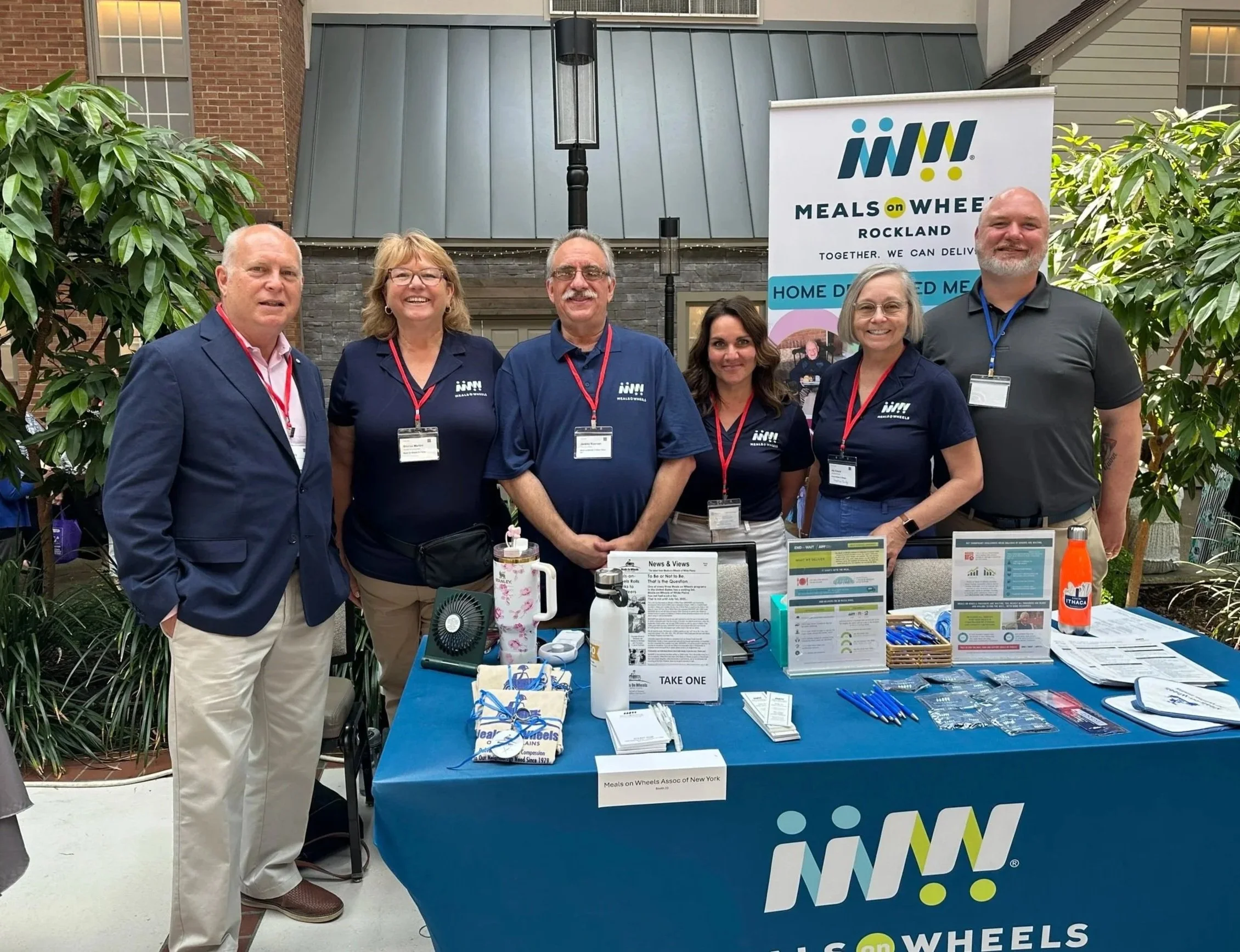 Meals on Wheels board members standing behind a table at an indoor event, promoting Meals on Wheels.