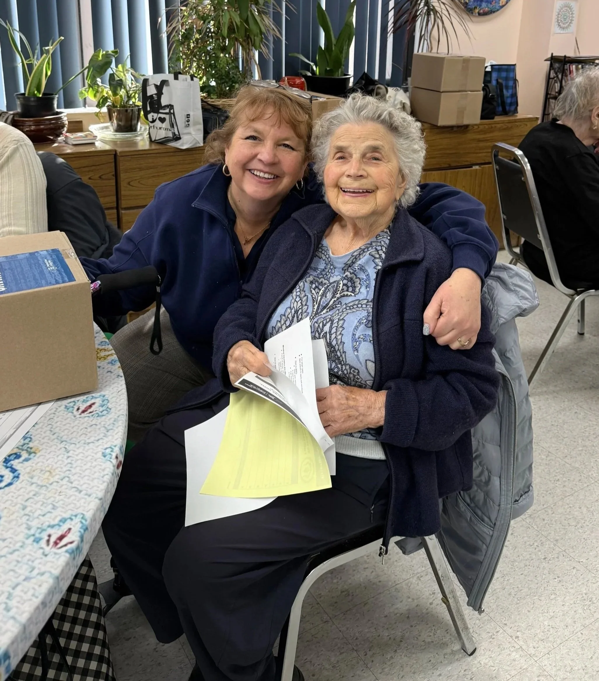 Two smiling women, one elderly with gray curly hair and the other middle-aged with short reddish hair, sitting closely together indoors. The elderly woman is holding papers, and the middle-aged woman has her arm around her. The background shows potted plants, a table with a colorful tablecloth, and various boxes and bags.