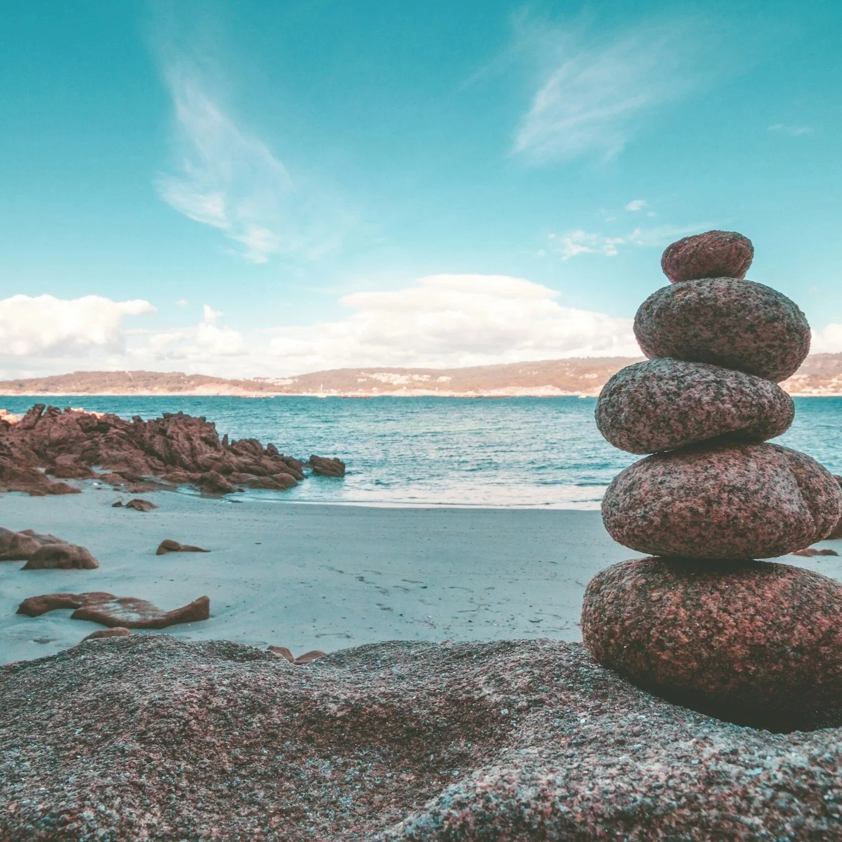 Stacked rocks arranged in a tower on a beach with rocks, water, distant land, and blue sky with clouds.