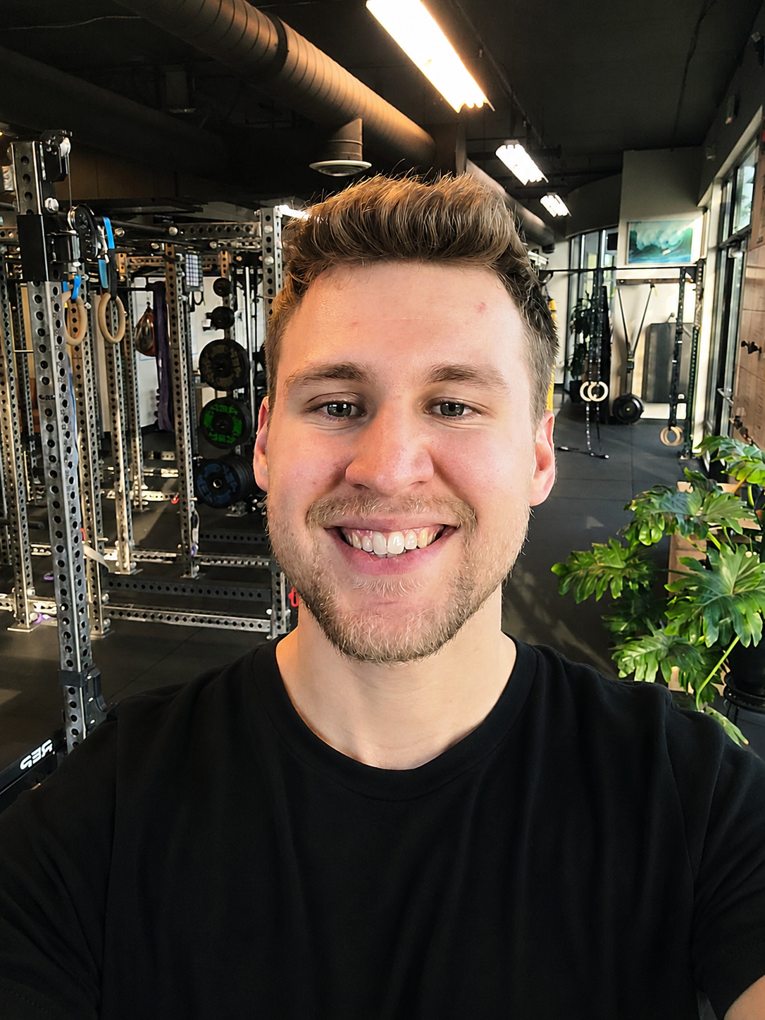 A smiling young man taking a selfie inside a gym, with workout equipment and green plant in the background.