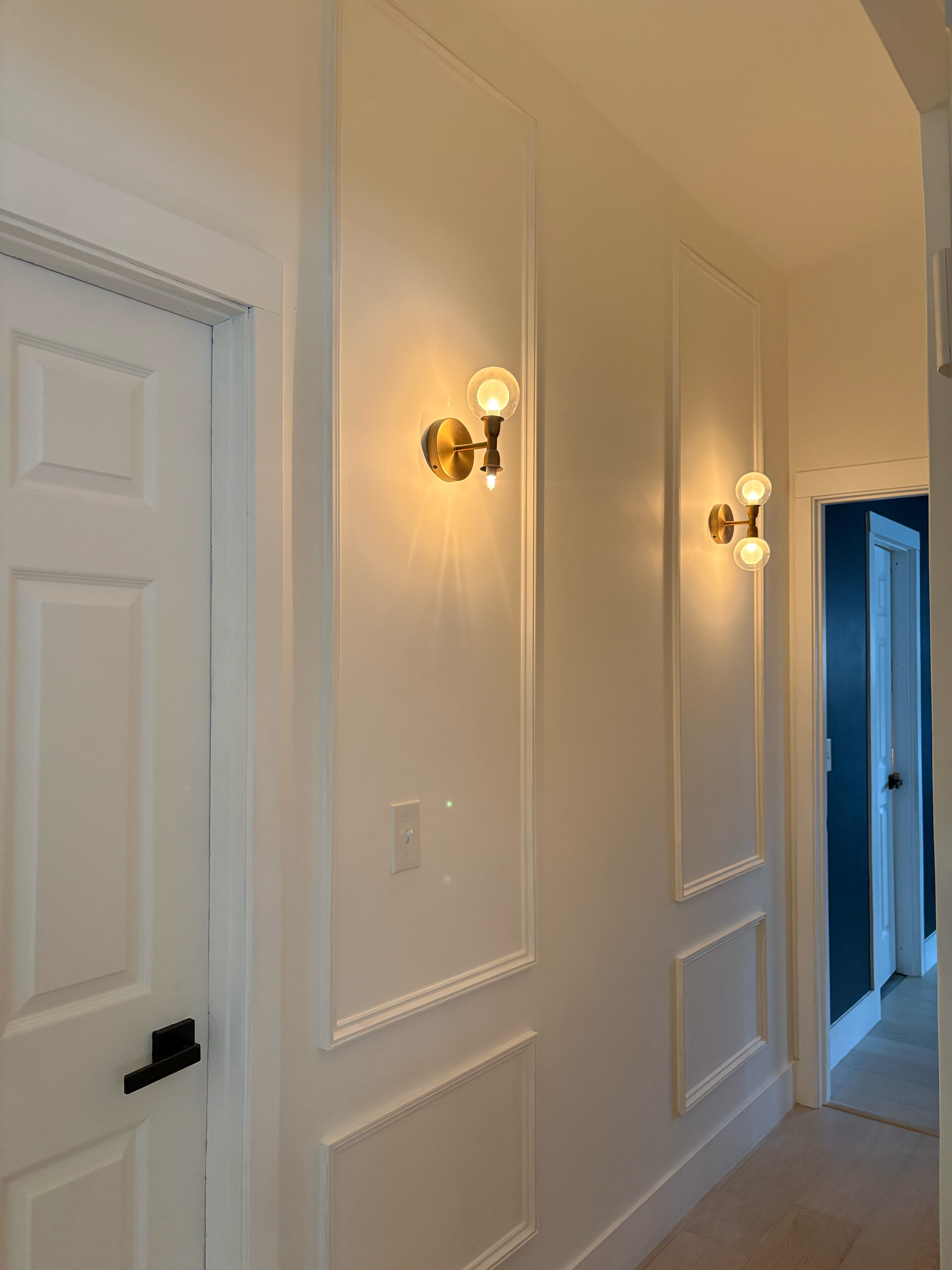Hallway with white paneled walls, two brass wall sconces with exposed bulbs, and an open doorway leading to a room with dark blue walls