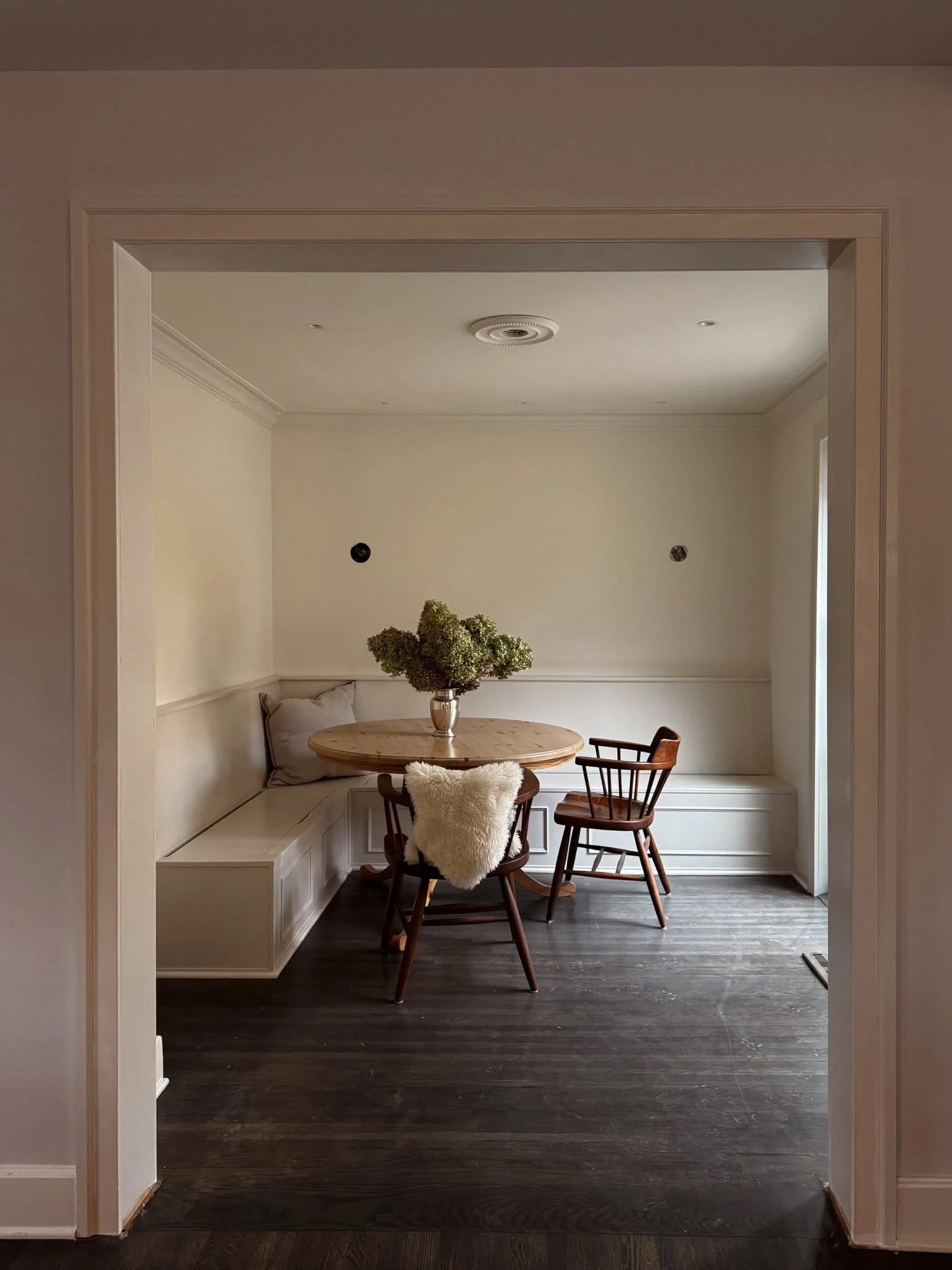 A dining nook with a round wooden table, a vase of green flowers, and three chairs, one with a fluffy white cushion, in a room with dark wood flooring and light-colored walls.