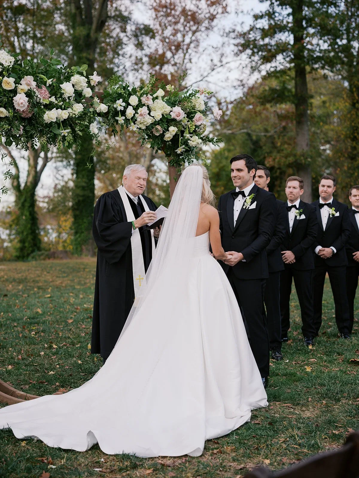 Bride and groom exchanging vows during an outdoor wedding ceremony with an officiant and groomsmen in the background, under a decorated floral arch.