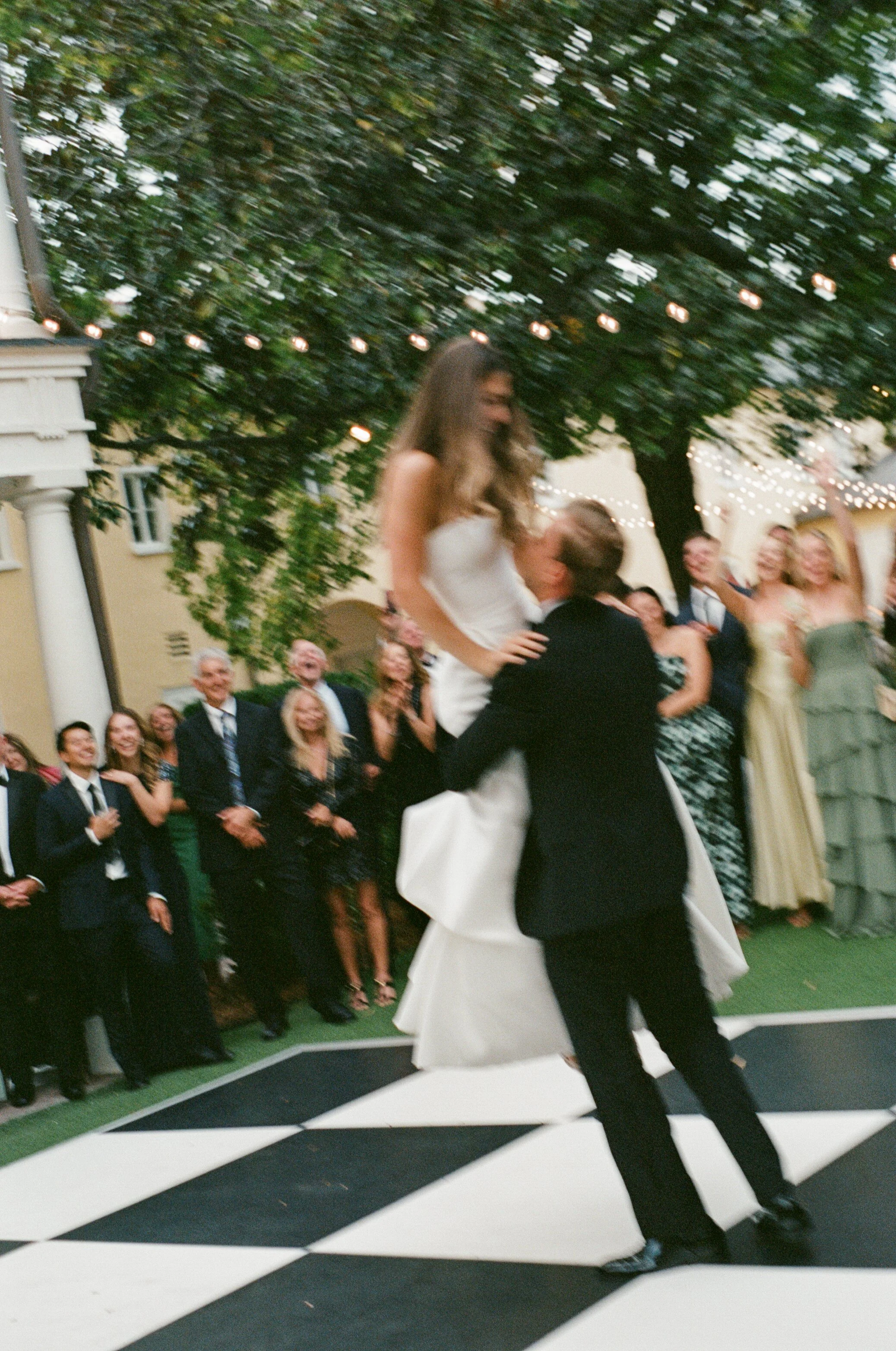 A wedding couple dancing on a checkered dance floor surrounded by guests at an outdoor reception with string lights and trees in the background.