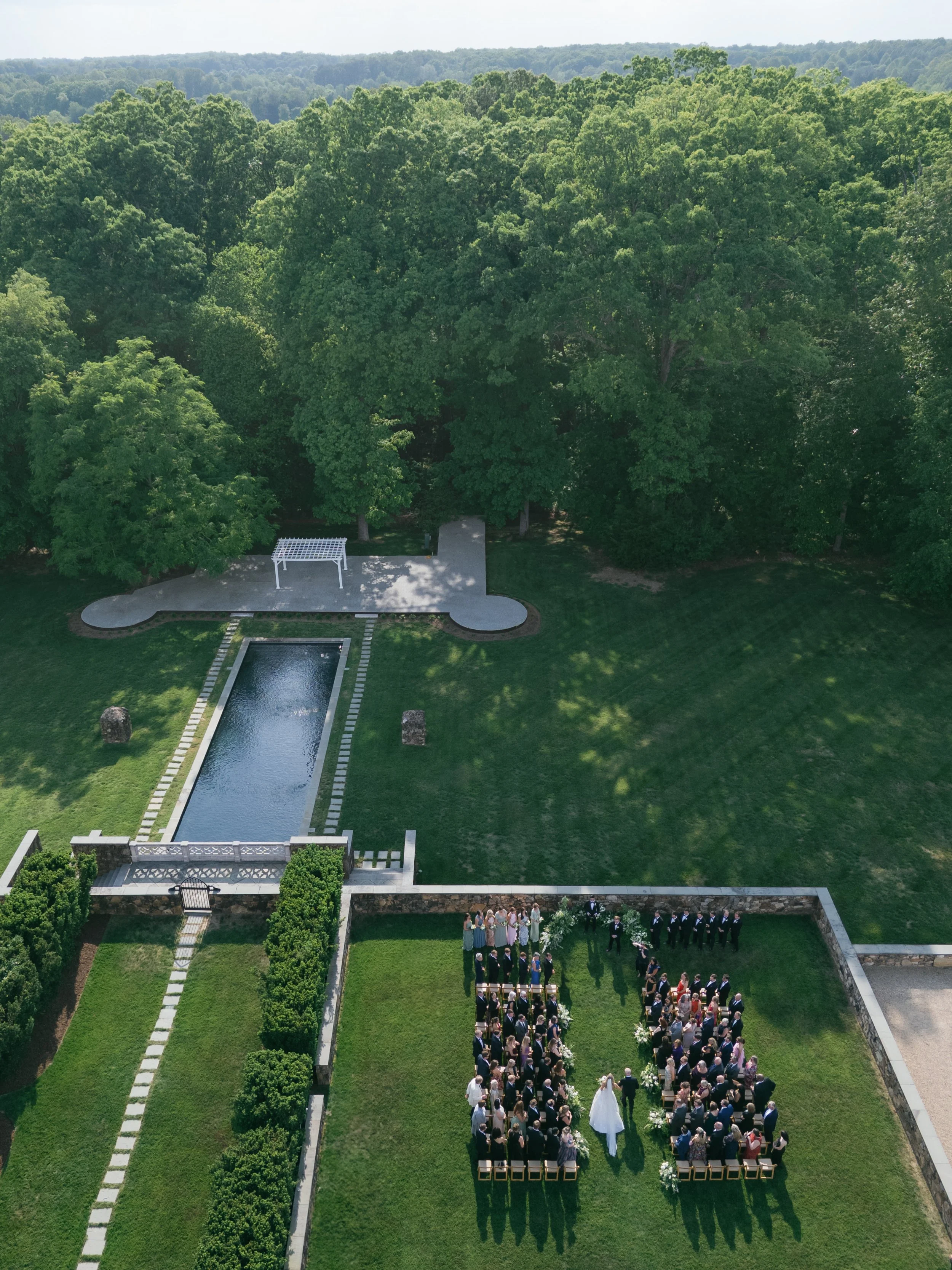 An outdoor wedding ceremony in a garden with guests seated around a bride and groom. The setting includes a rectangular pool, lush green trees, and a spacious lawn.