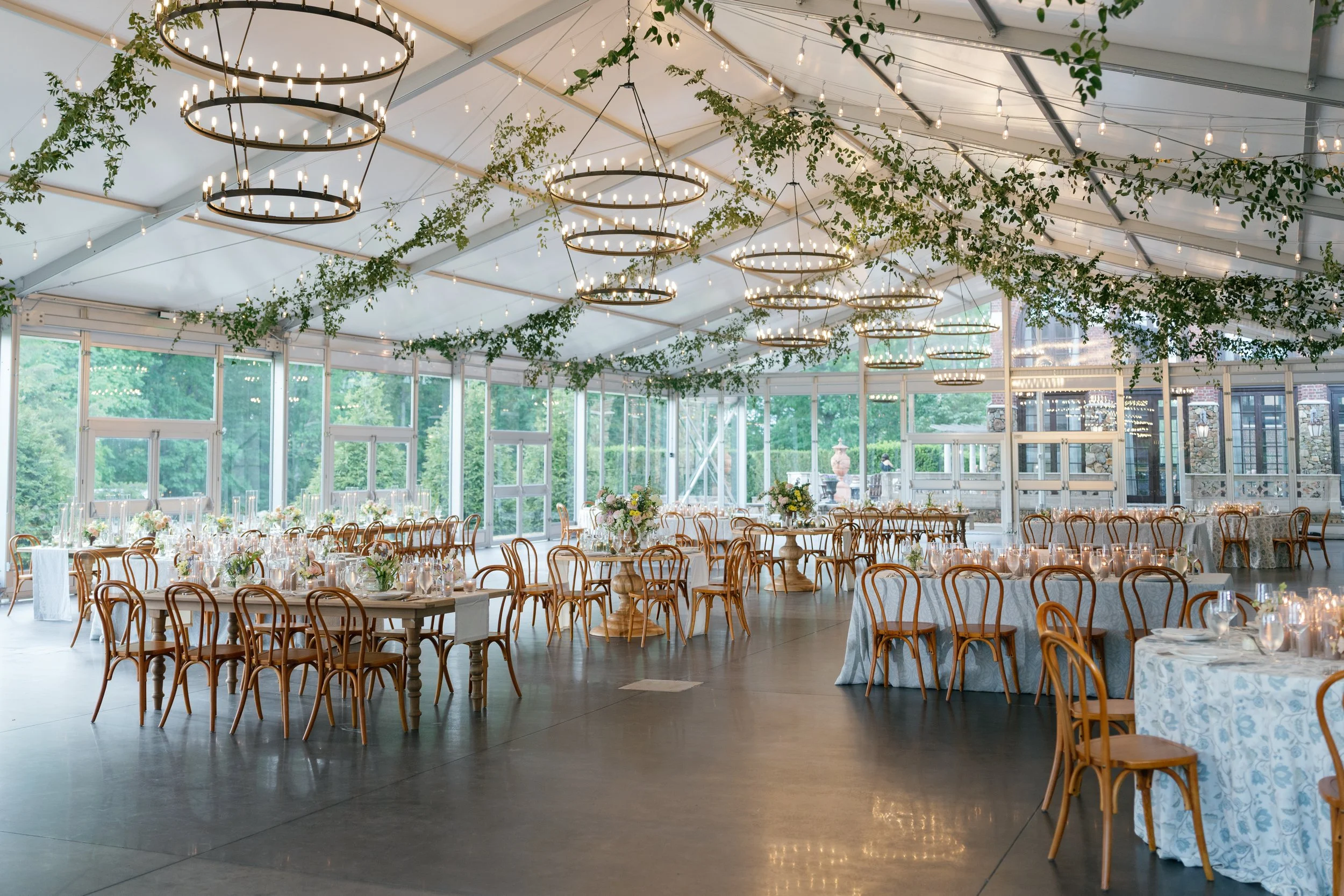 Indoor wedding reception area with round and rectangular tables, wooden chairs, floral centerpieces, string lights, and greenery hanging from the ceiling, with large windows showing a green outdoor landscape.