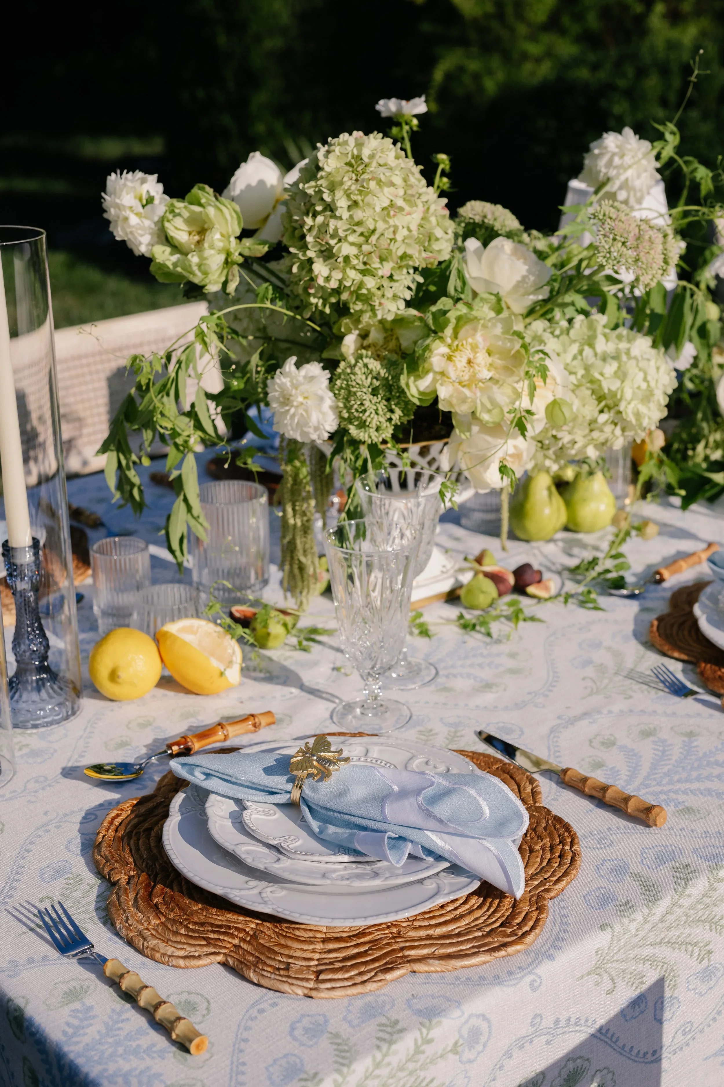 Elegant outdoor table setting with white flowers centerpiece, green pears, lemons, cutlery with bamboo handles, glassware, and a decorative tablecloth.