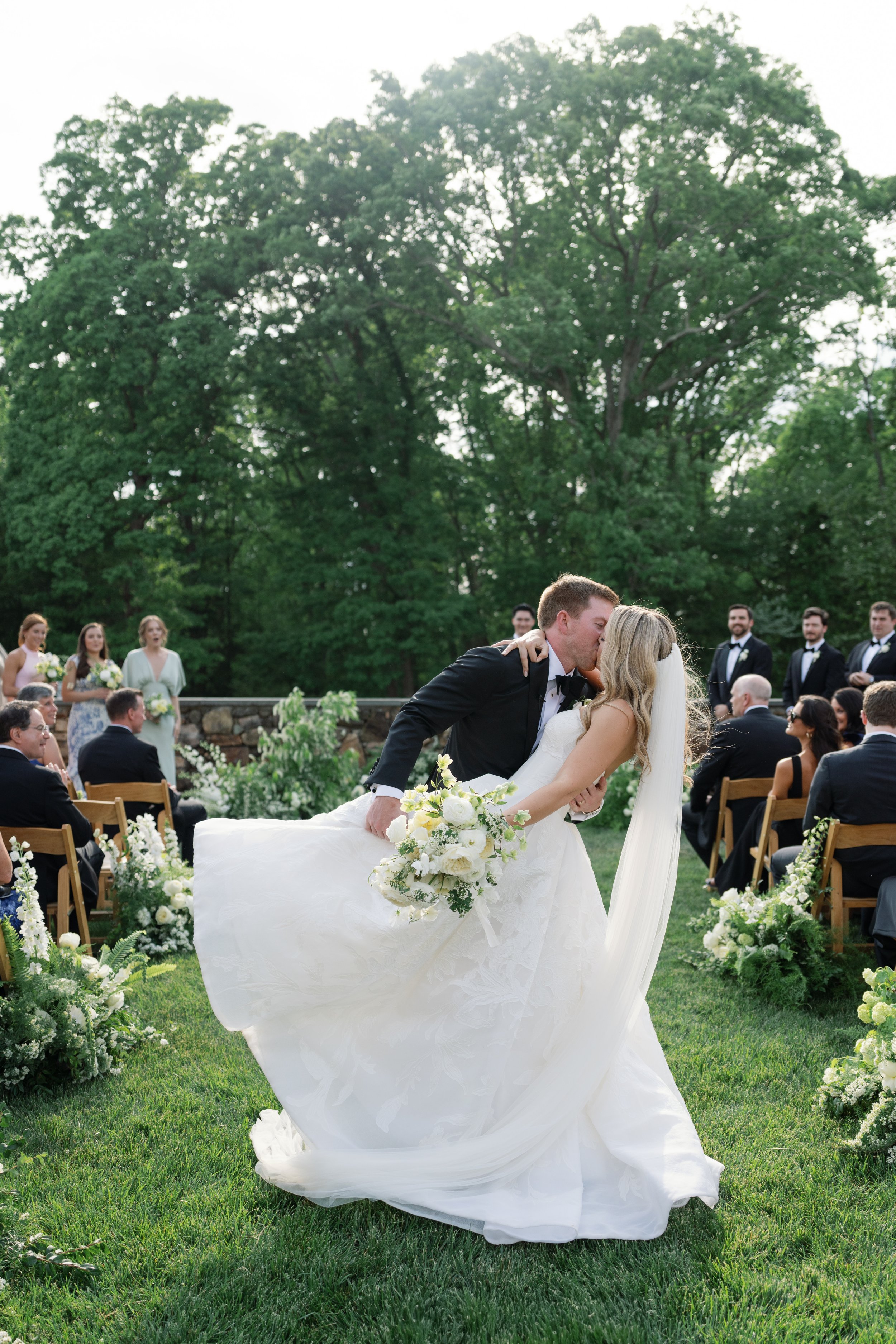 A newlywed couple shares a kiss during an outdoor wedding ceremony, with guests seated on either side and green trees in the background.