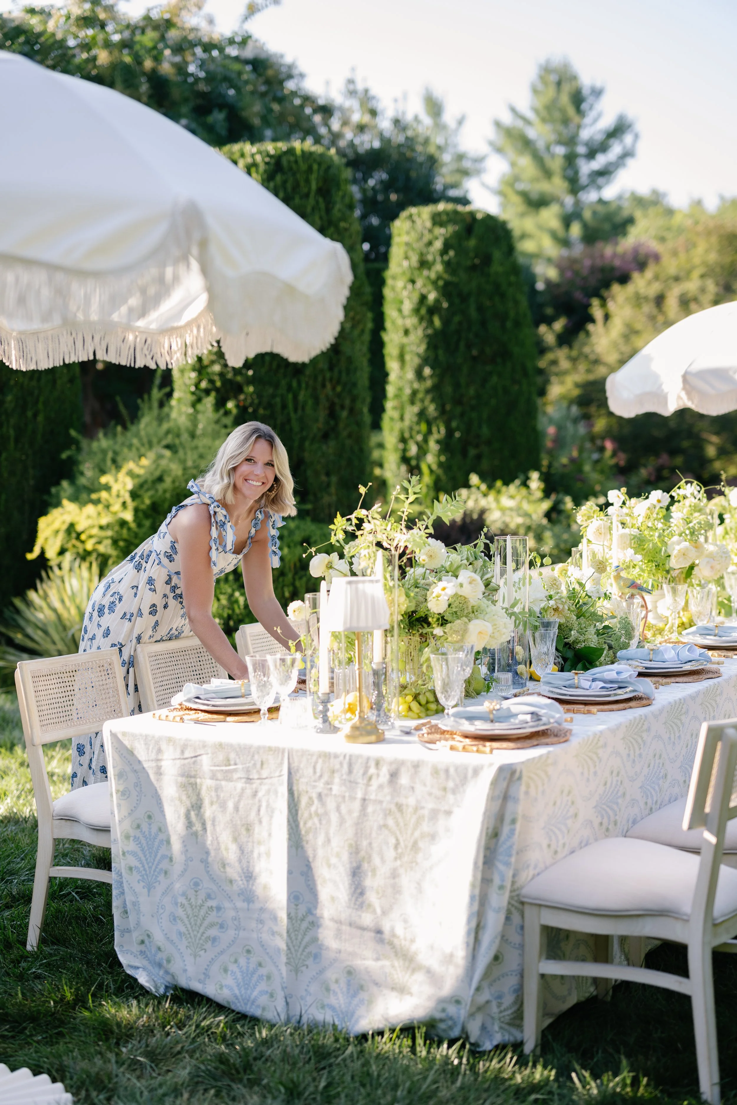 A woman in a floral dress arranging a long outdoor table set for a meal, with white umbrellas providing shade, surrounded by green trees and bushes.