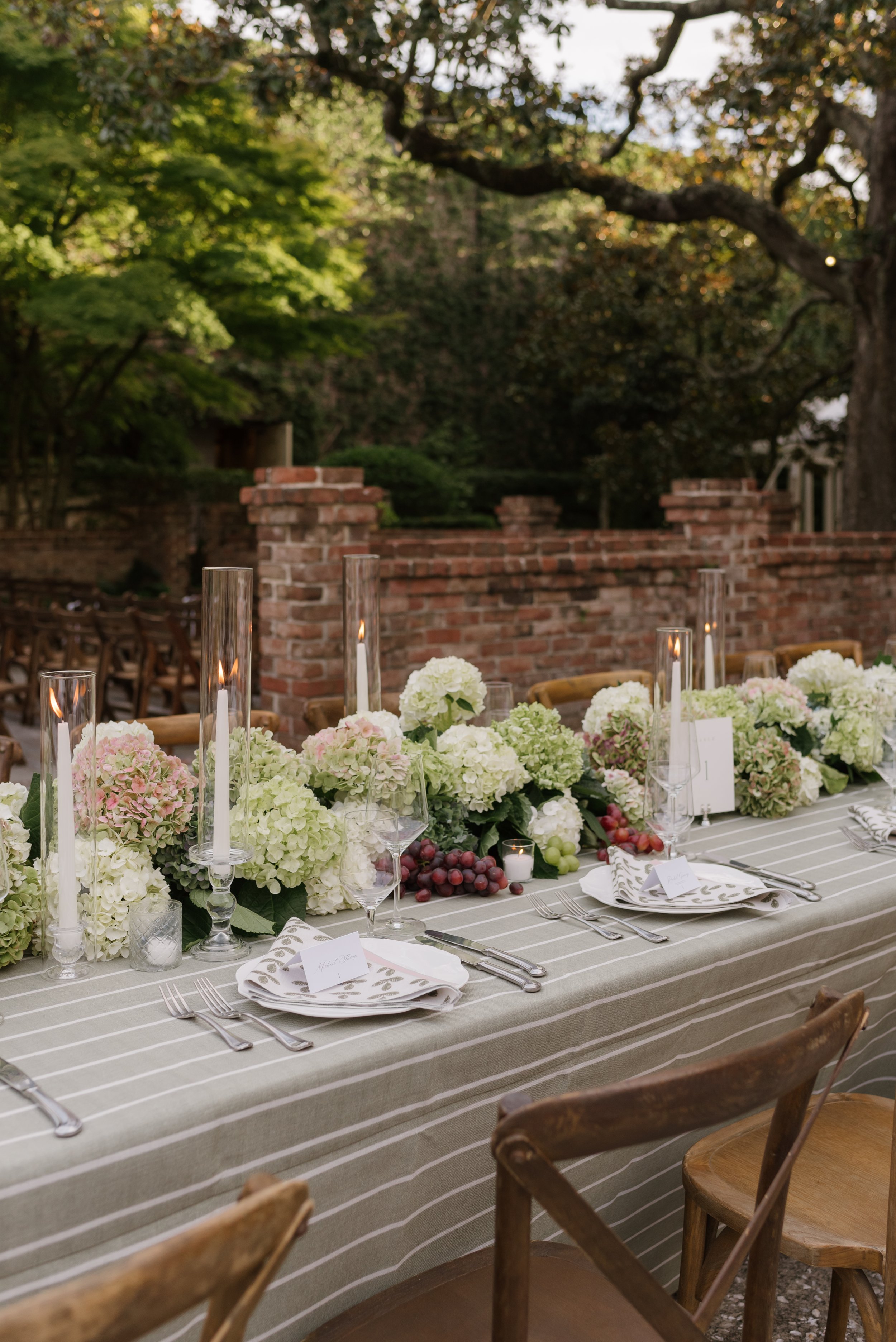 An outdoor dining table decorated with white hydrangeas, pink and green flowers, grapes, and tall glass candle holders with lit candles, set for a formal meal in front of a brick wall and lush green trees.