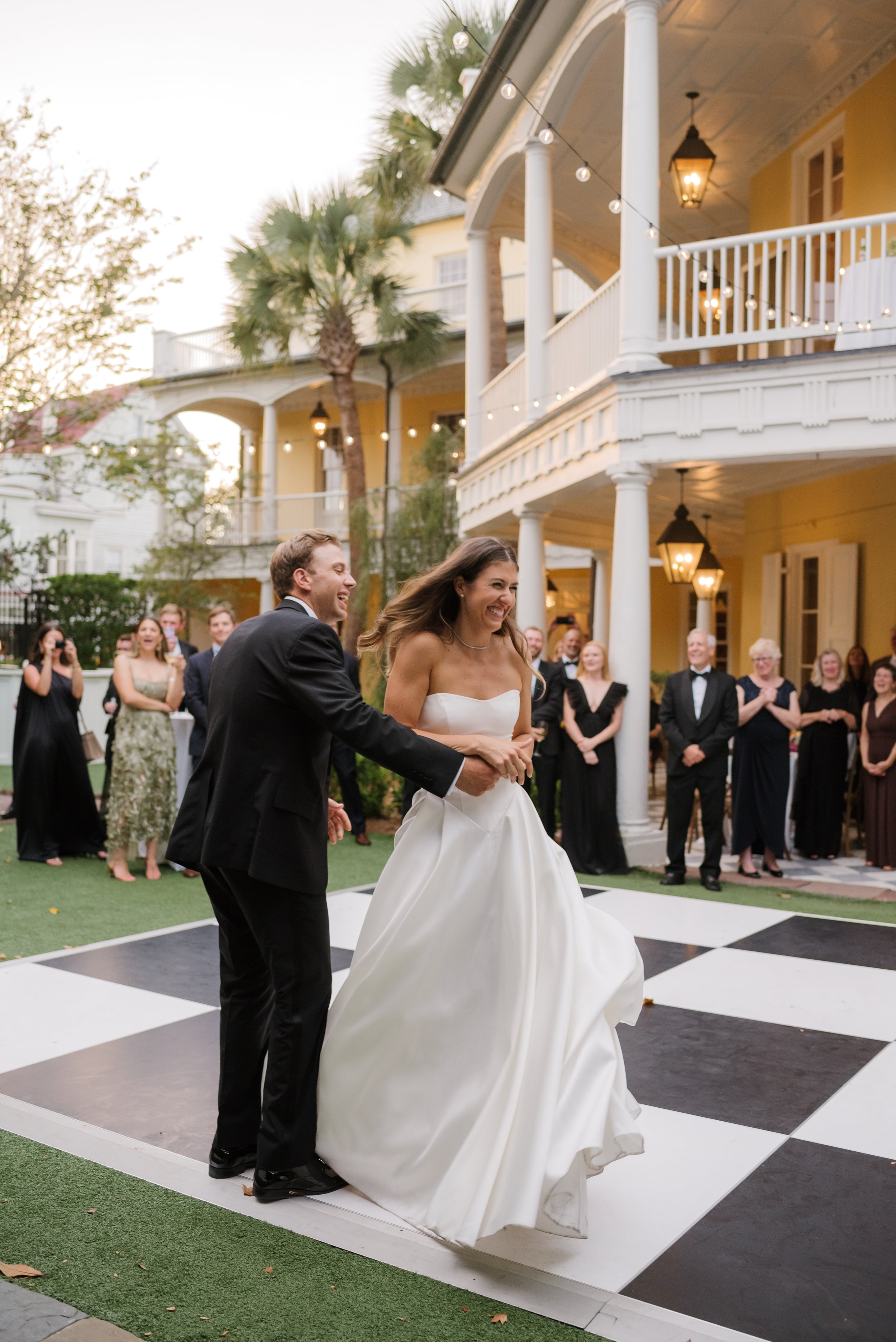 A bride and groom dancing on a life-sized chessboard outside a yellow house, surrounded by wedding guests watching and smiling.