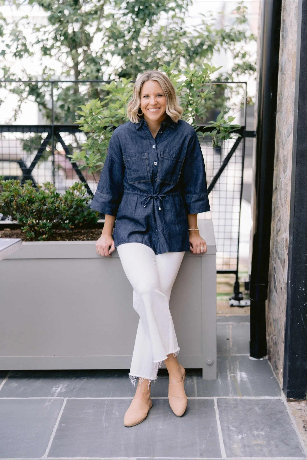 A woman with blonde hair wearing a denim dress with a drawstring waist, white frayed hem jeans, and beige shoes, standing outdoors in front of plants and smiling.