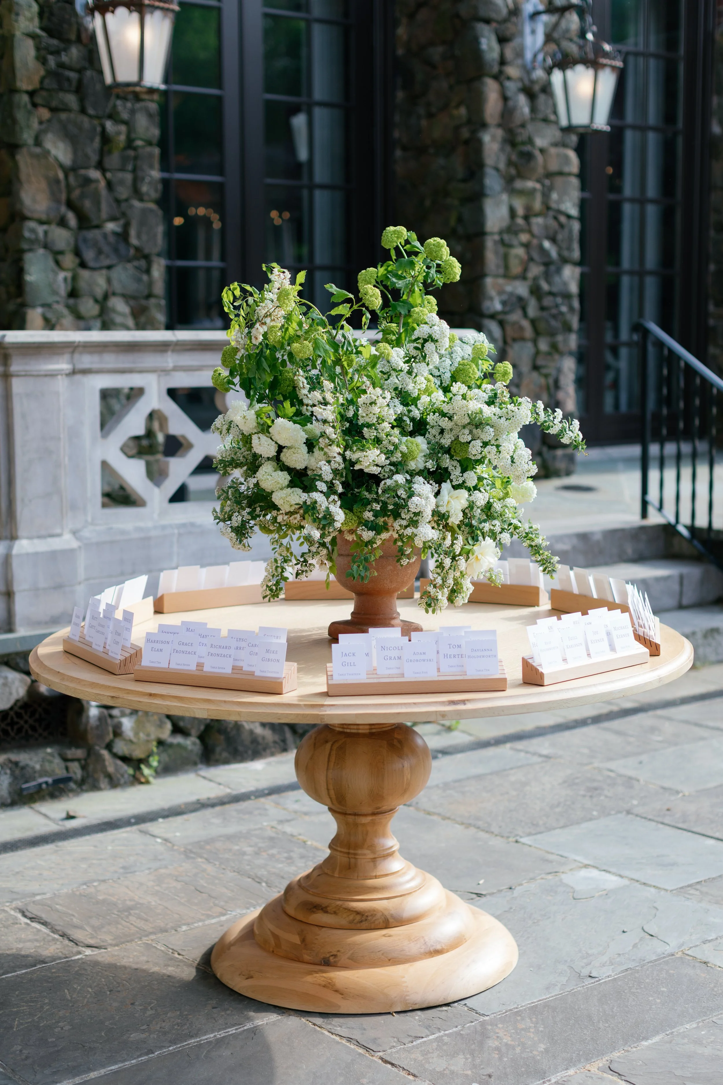 A floral arrangement of white and green flowers on a wooden table, with name tags arranged in outer trays surrounding the centerpiece, outside a stone building with black-framed windows.