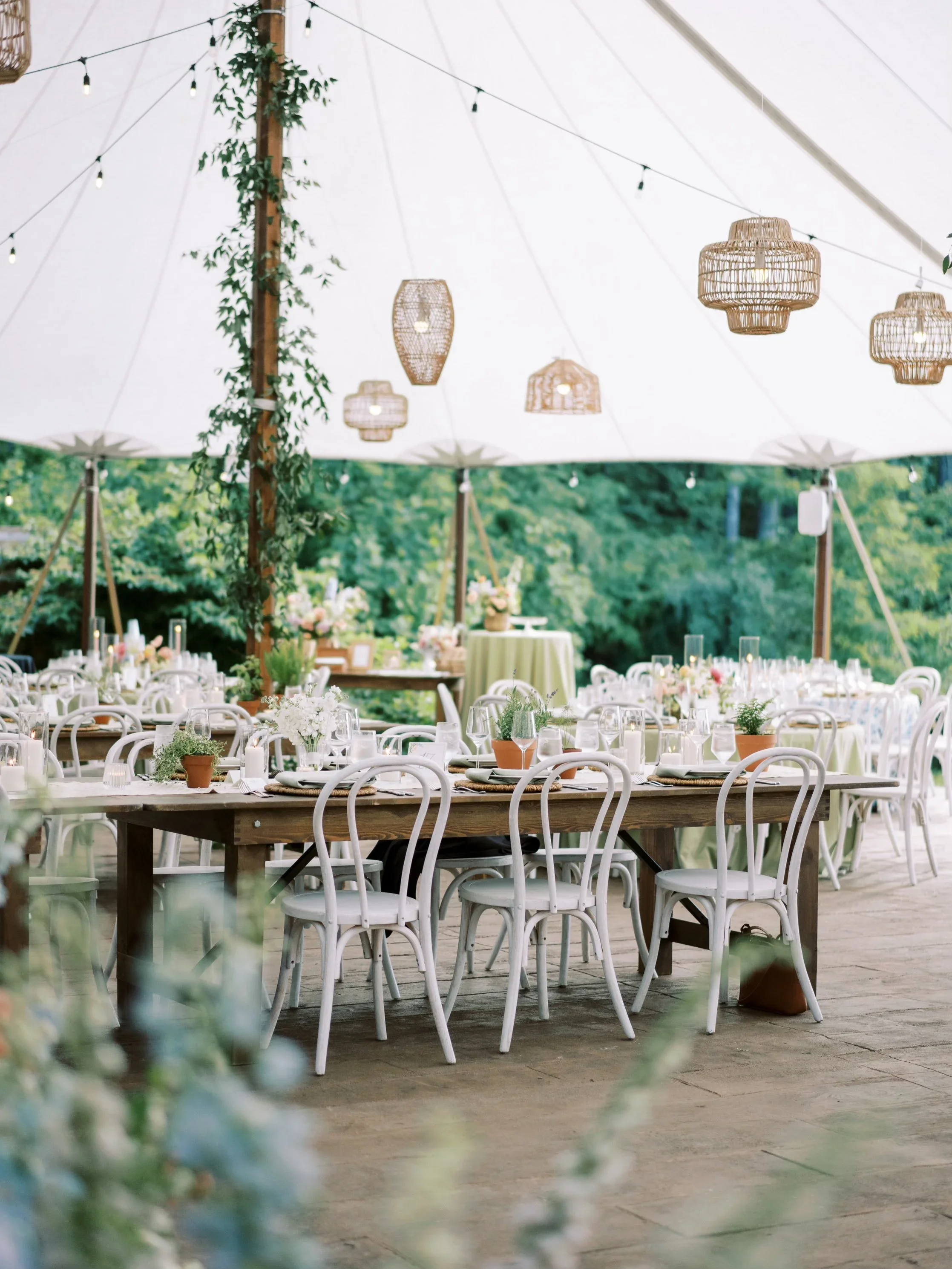 Outdoor event space with decorated tables and chairs under a large white tent with hanging wicker lamps and string lights, surrounded by green trees.
