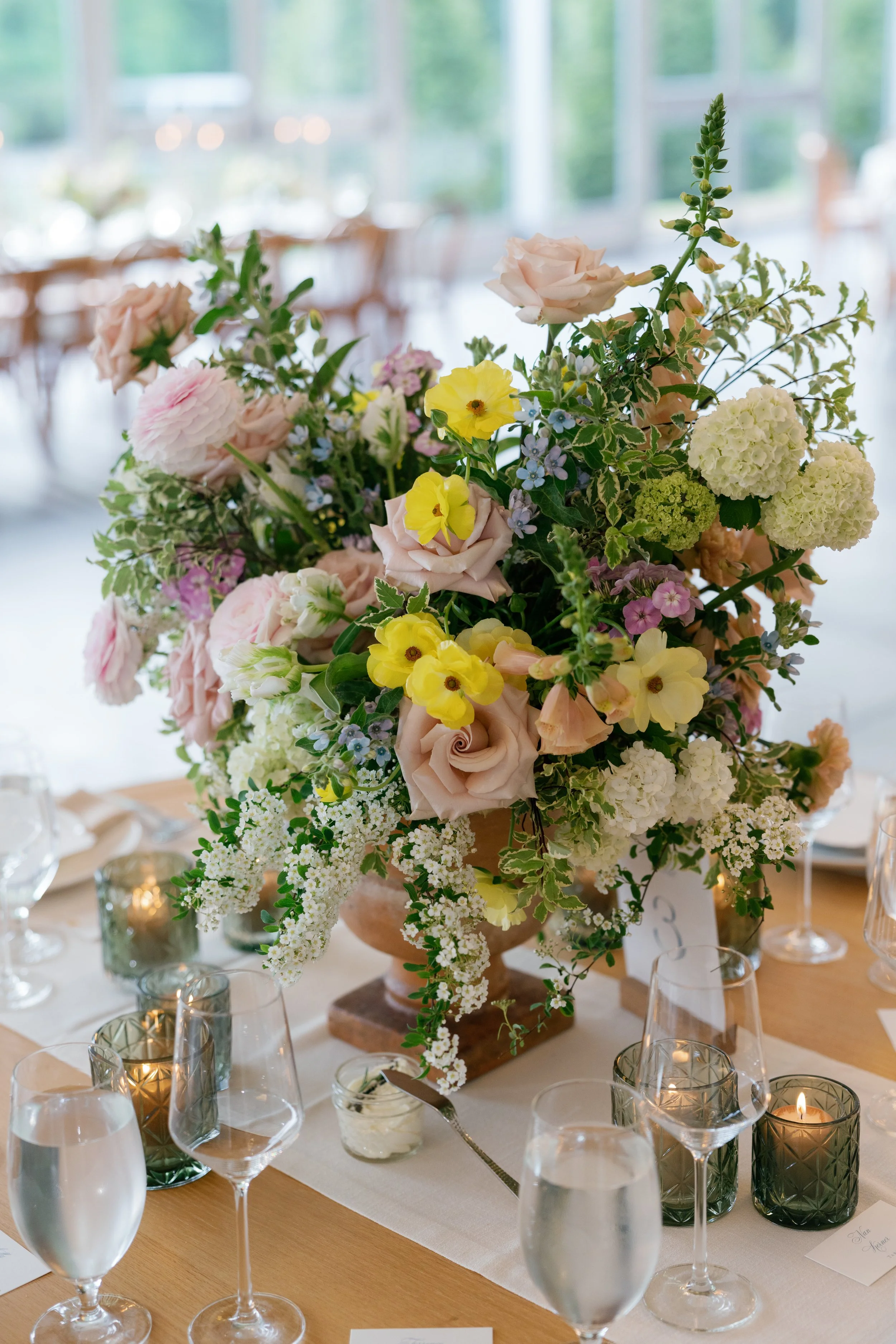 A large floral centerpiece with roses, hydrangeas, and other flowers on a dining table, surrounded by glasses and candles.