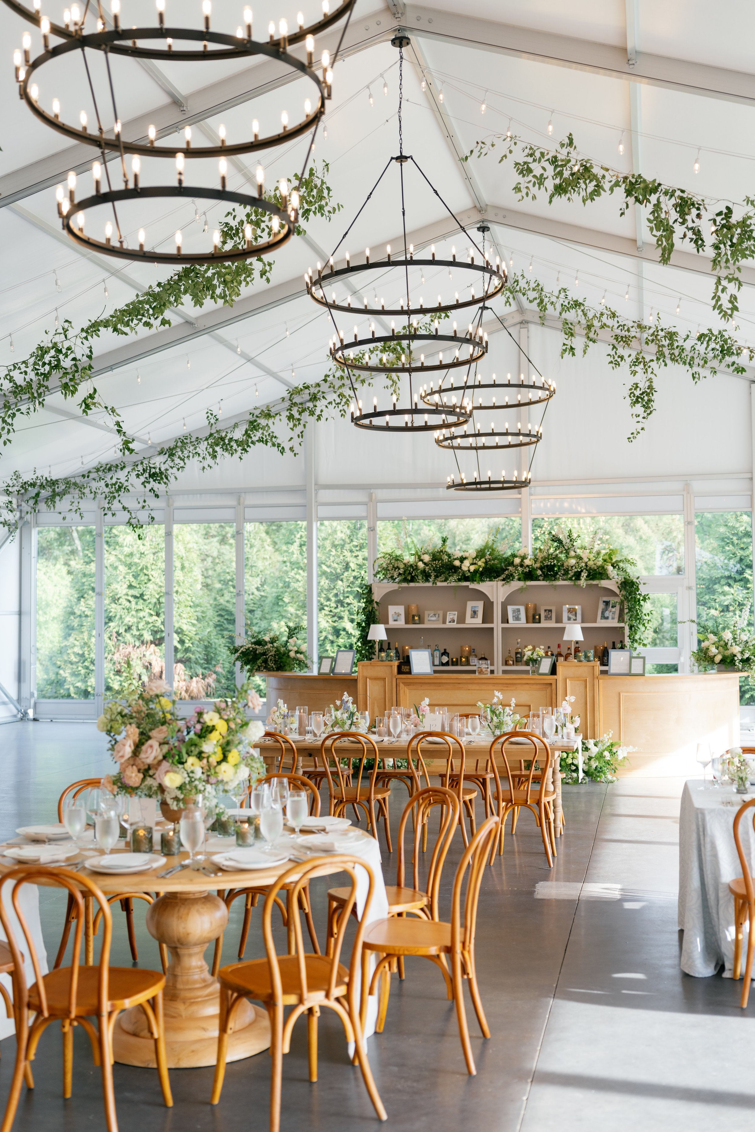 Elegant indoor wedding reception with a high ceiling decorated with hanging greenery and chandelier lights, floral arrangements on round and rectangular tables, and a bar area in the background, all illuminated by natural light from large windows.