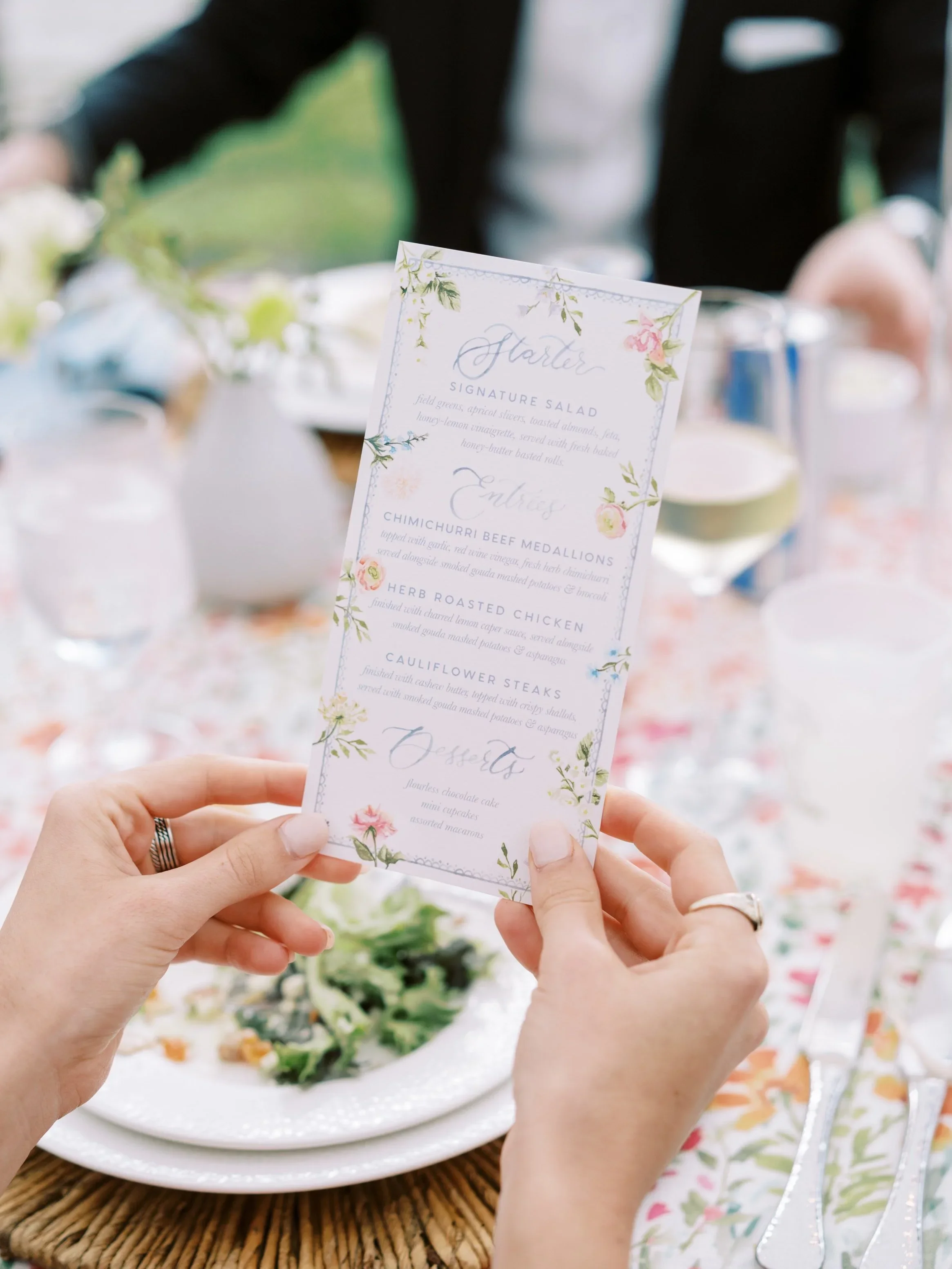 Person holding a wedding menu featuring floral details, with a plate of food, wine glass, and tableware set on a decorated wedding table in the background.