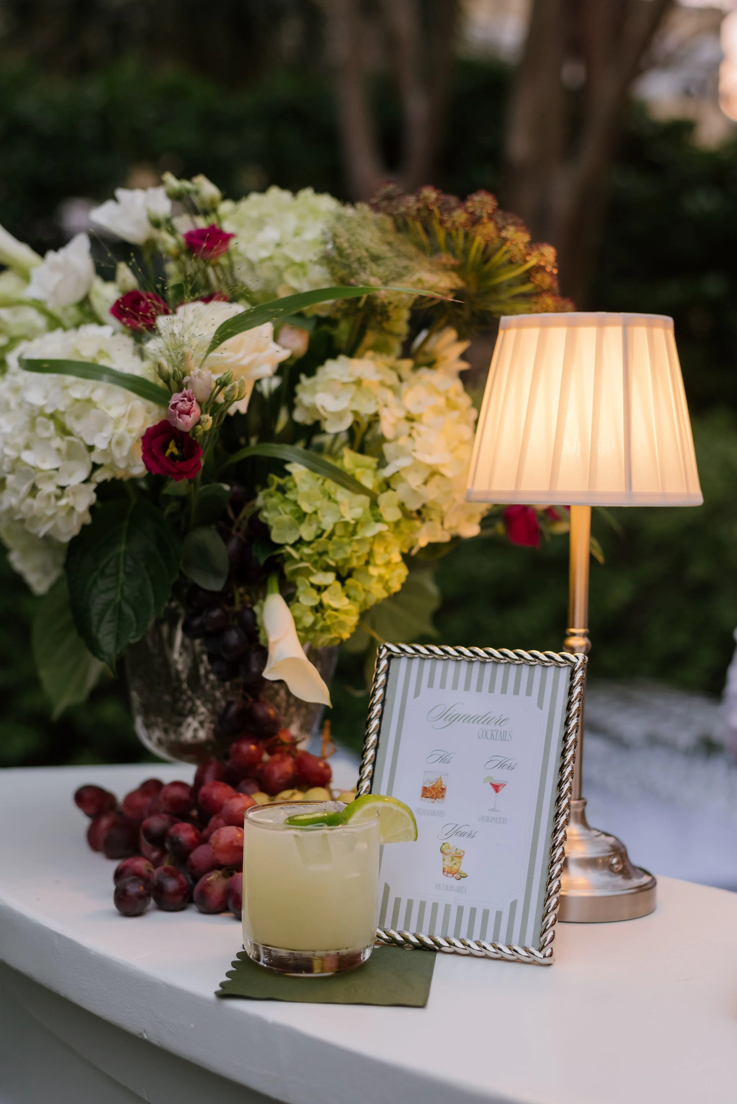 A floral arrangement with white, pink, and purple flowers, and a pitcher of lemonade garnished with lime, on a table next to a small lamp. There is a framed sign listing signature cocktails.
