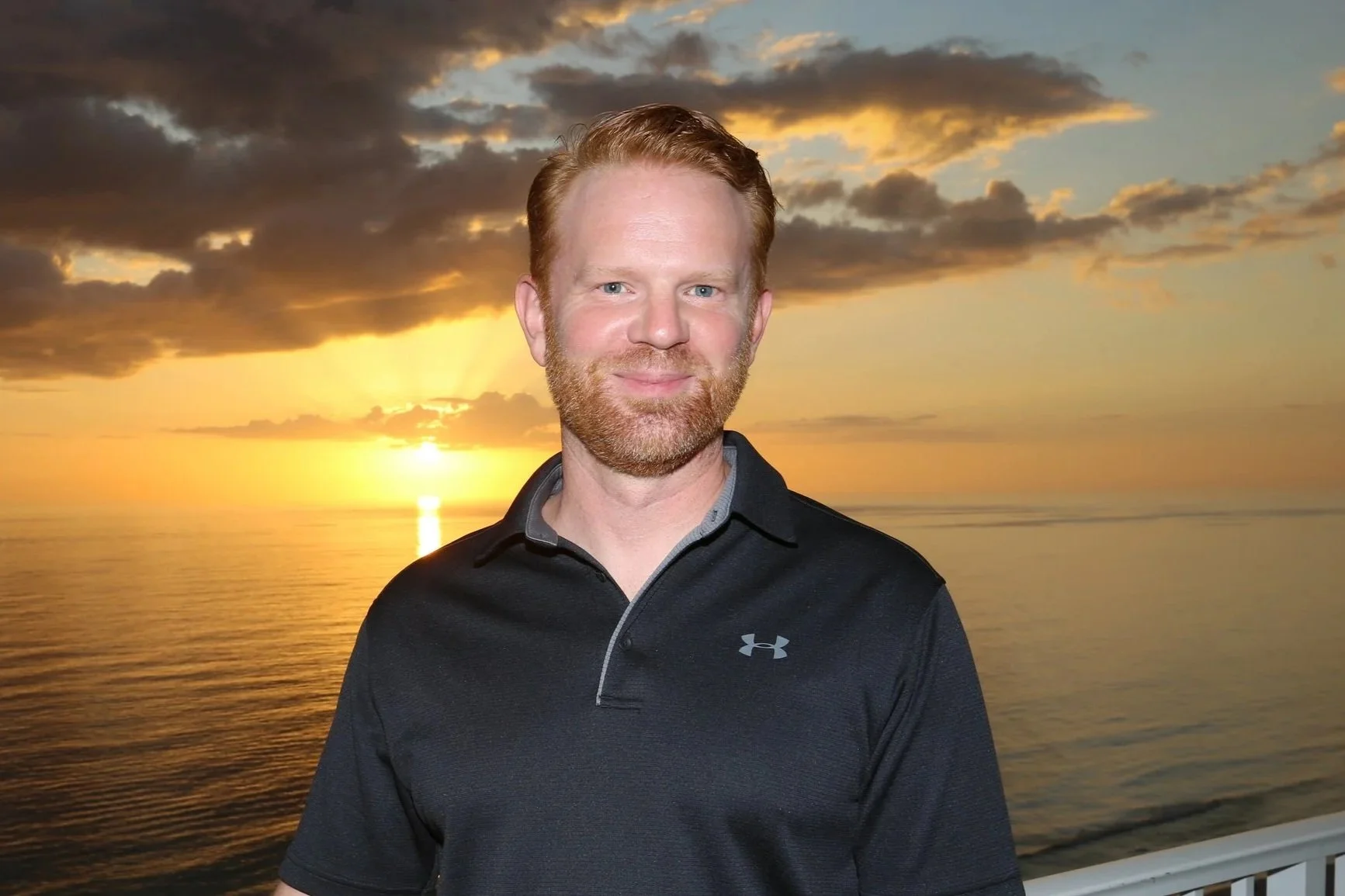 A man with red hair and a beard standing on a balcony near the ocean during sunset, wearing a black Under Armour polo shirt.