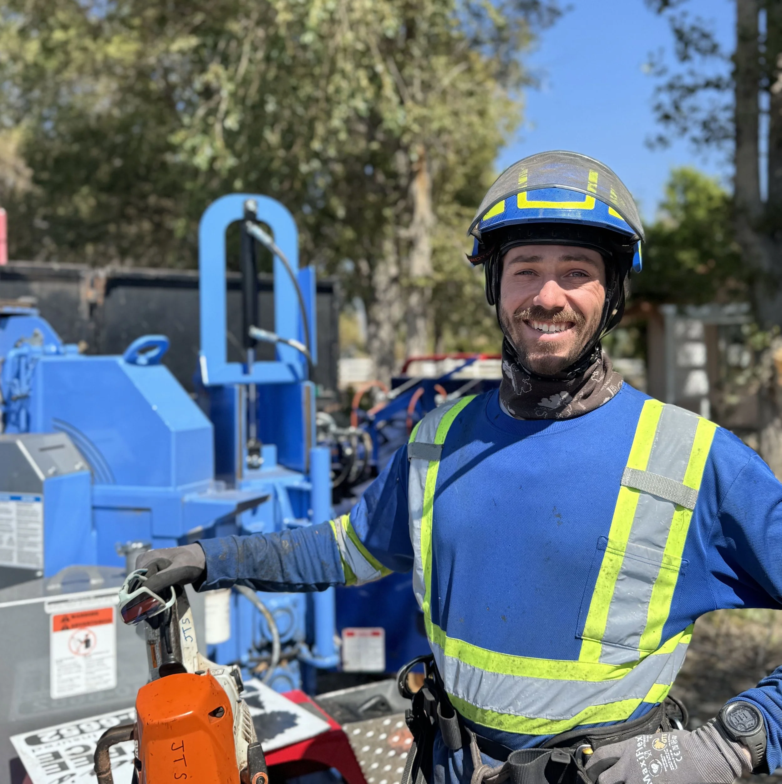 A smiling worker in safety gear standing outdoors near machinery, holding a chainsaw.