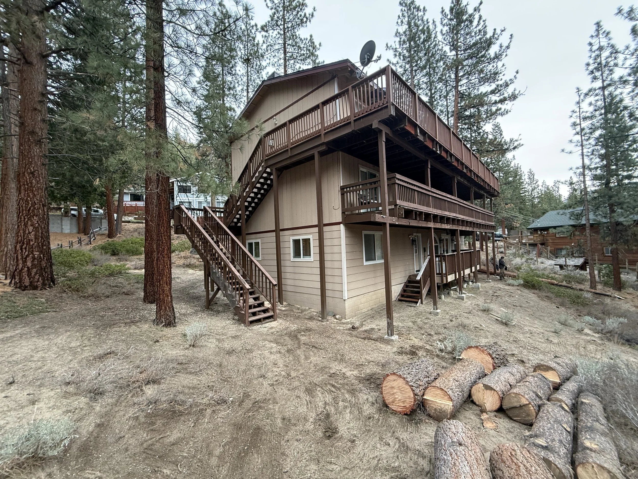 A beige multi-story house with brown wooden decks and stairs, surrounded by tall pine trees, with logs stacked on the ground nearby.