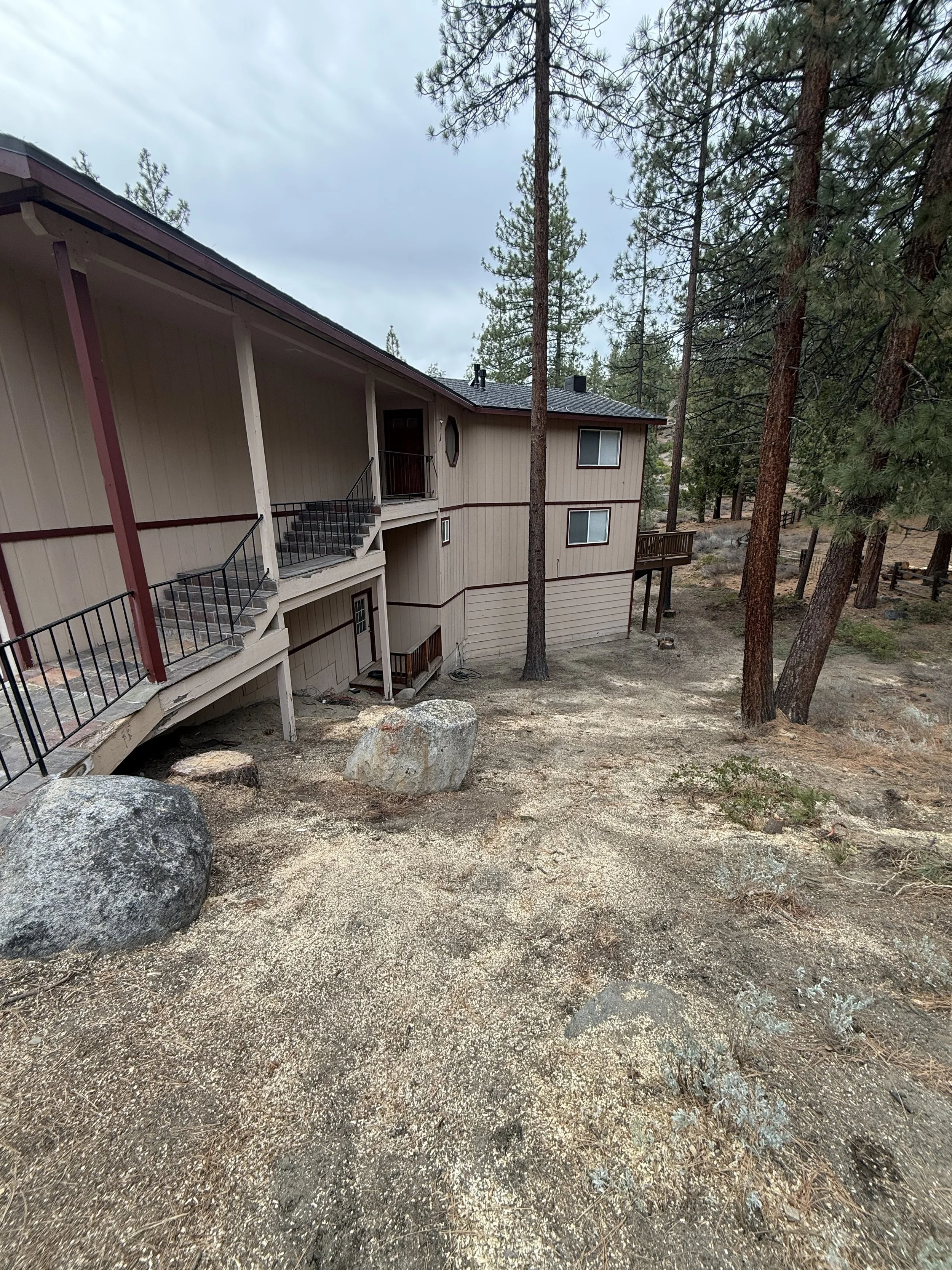 A multi-story beige house with a deck and stairs, surrounded by tall pine trees and rocky terrain under a cloudy sky.