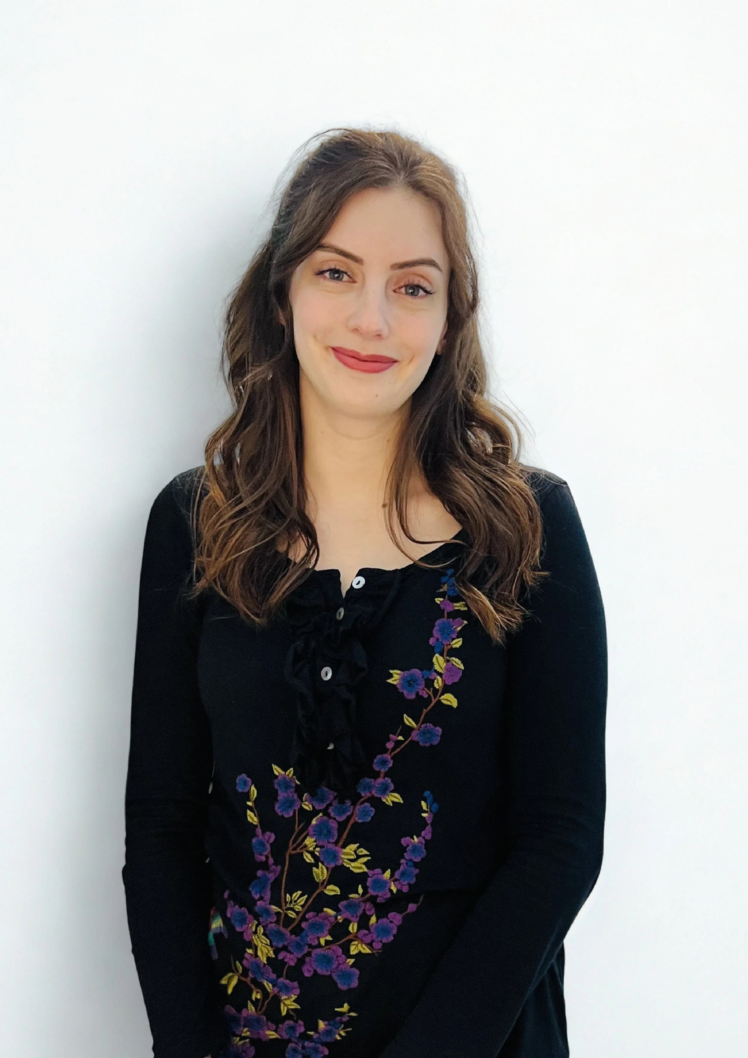 A young woman with shoulder-length brown hair, wearing a black top with purple and yellow floral embroidery, standing against a plain white wall.