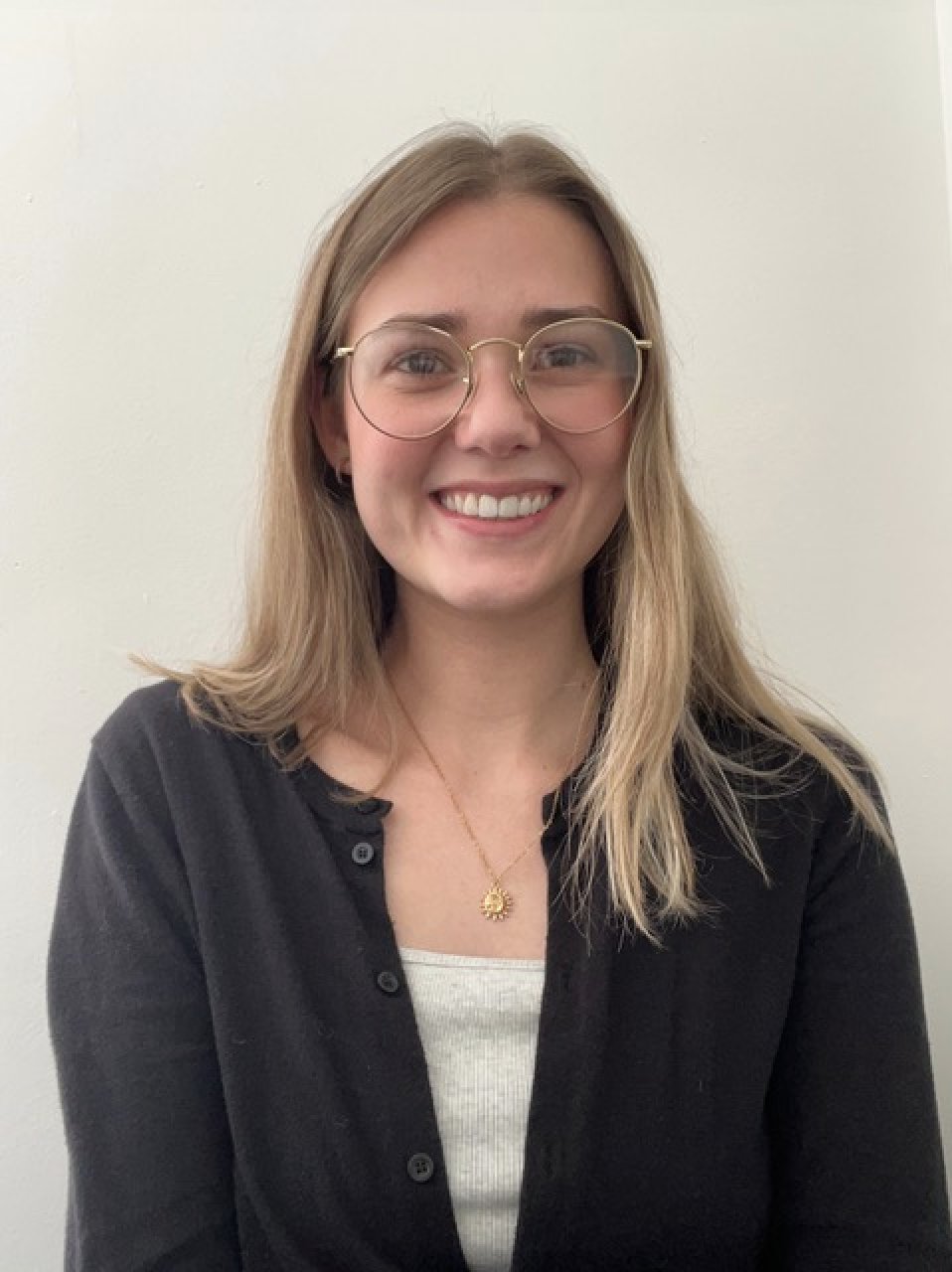 A young woman with blonde hair, glasses, and a big smile, wearing a black cardigan, white shirt, and a gold necklace, standing against a plain white wall.
