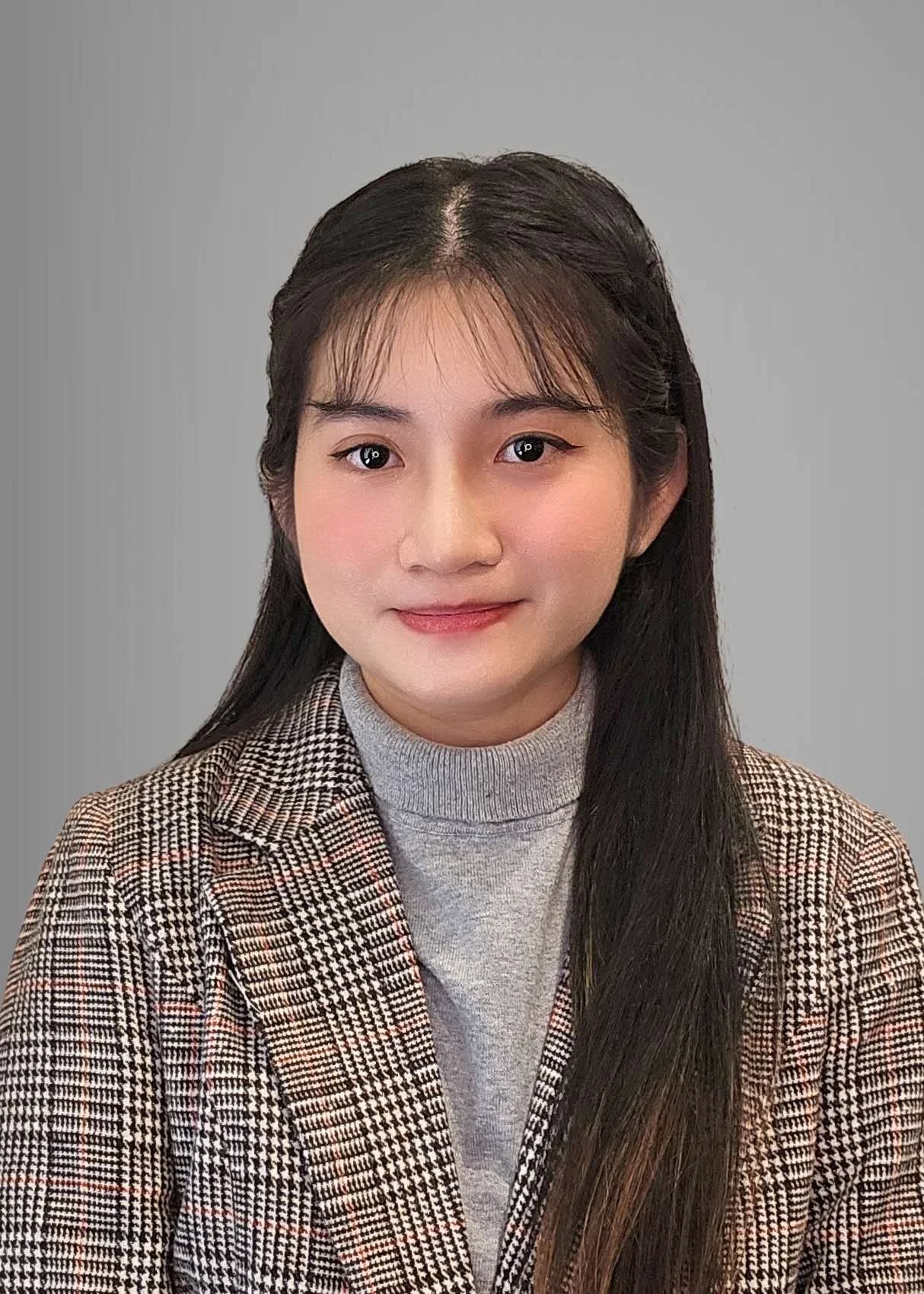 Portrait of a young Asian woman with long dark hair, wearing a light gray turtleneck and a houndstooth blazer, standing against a neutral background.