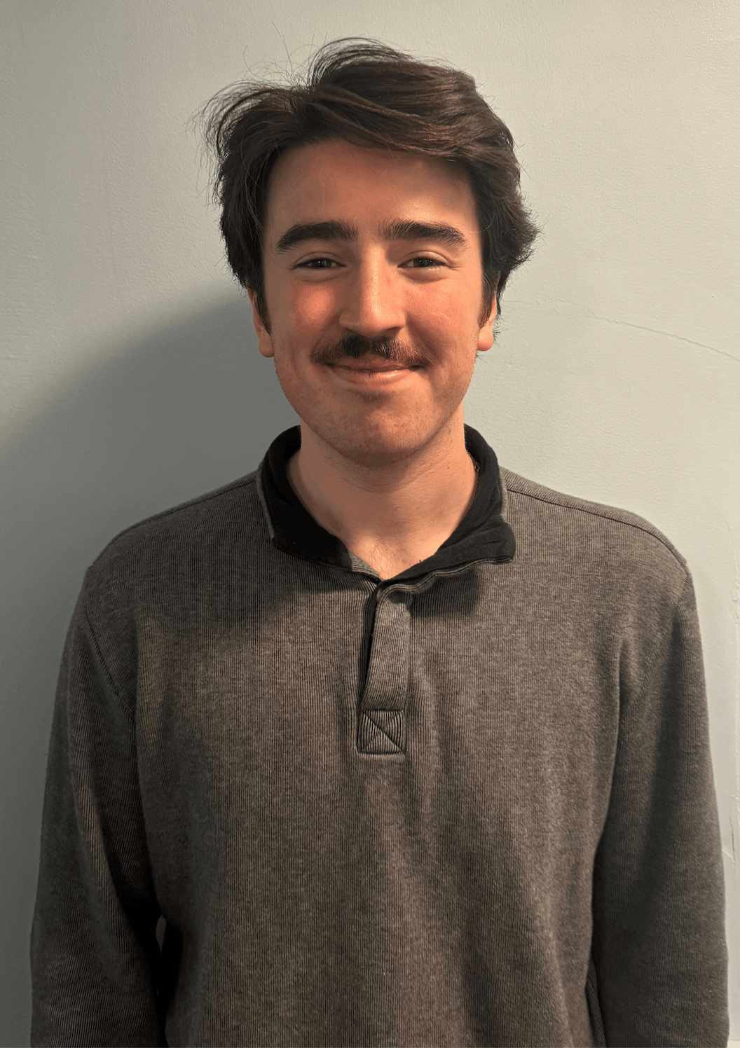 A young man with dark hair and a mustache, smiling, wearing a dark gray sweater, standing in front of a light-colored wall.