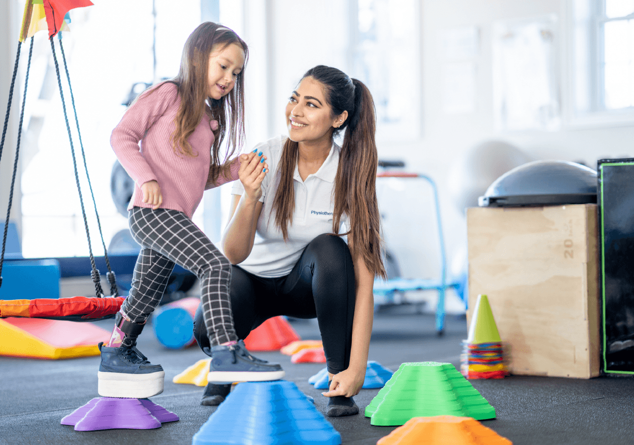 A woman assisting a young girl in an indoor physical therapy or exercise session, with colorful cones and balance equipment.