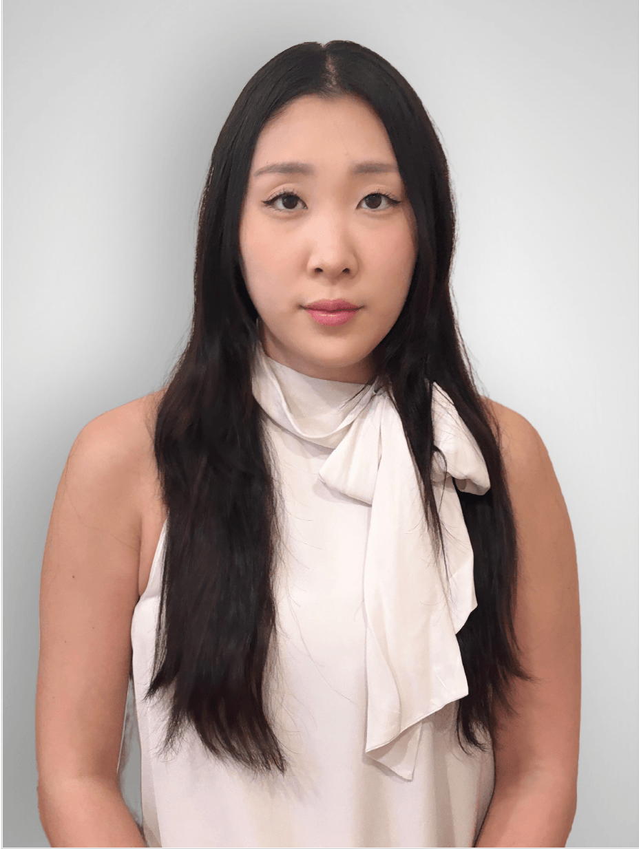 A young woman with long dark hair, wearing a sleeveless white top with a bow, standing against a plain light-colored background.