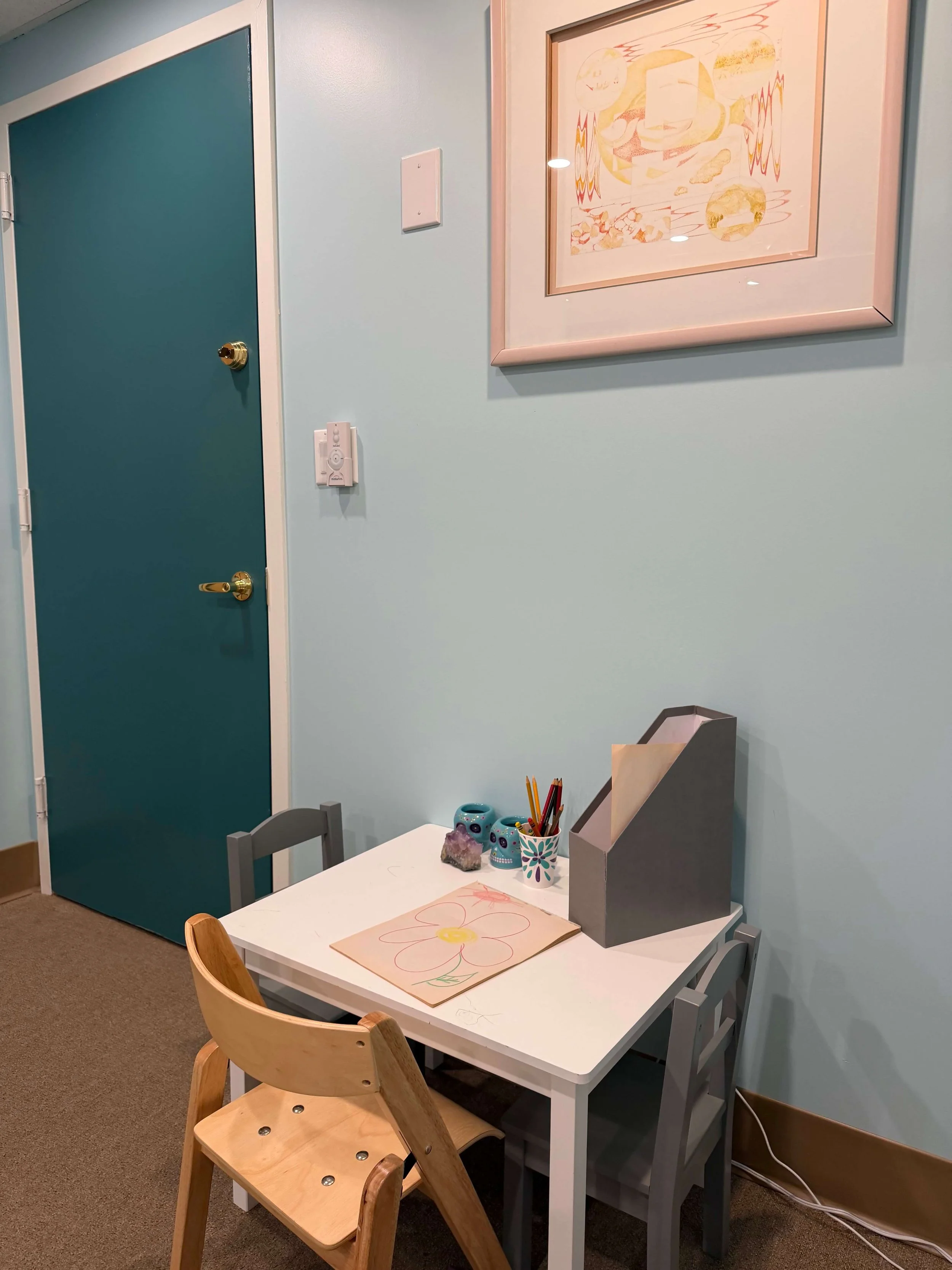 A small table with two chairs, one wooden and one grey, placed against a blue wall. The table has children's coloring pages, a container with coloring pencils, and decorative skull-shaped containers. A framed abstract painting hangs on the wall above