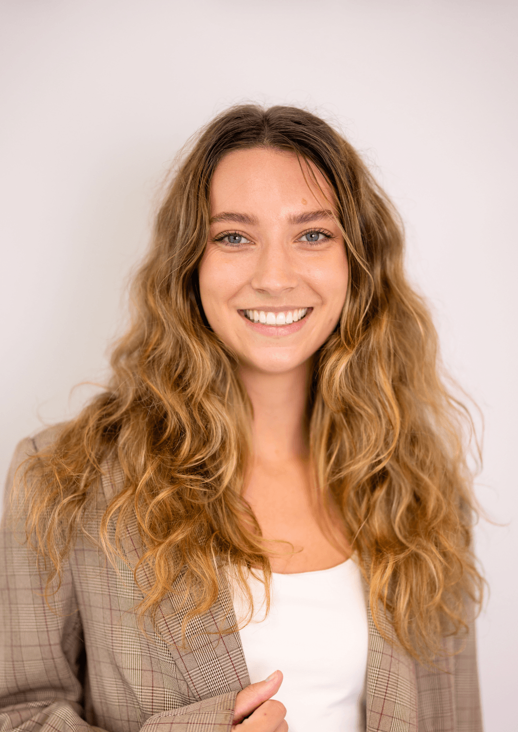 A smiling woman with long, wavy, light brown hair, wearing a tan plaid blazer over a white shirt, standing against a plain white background.