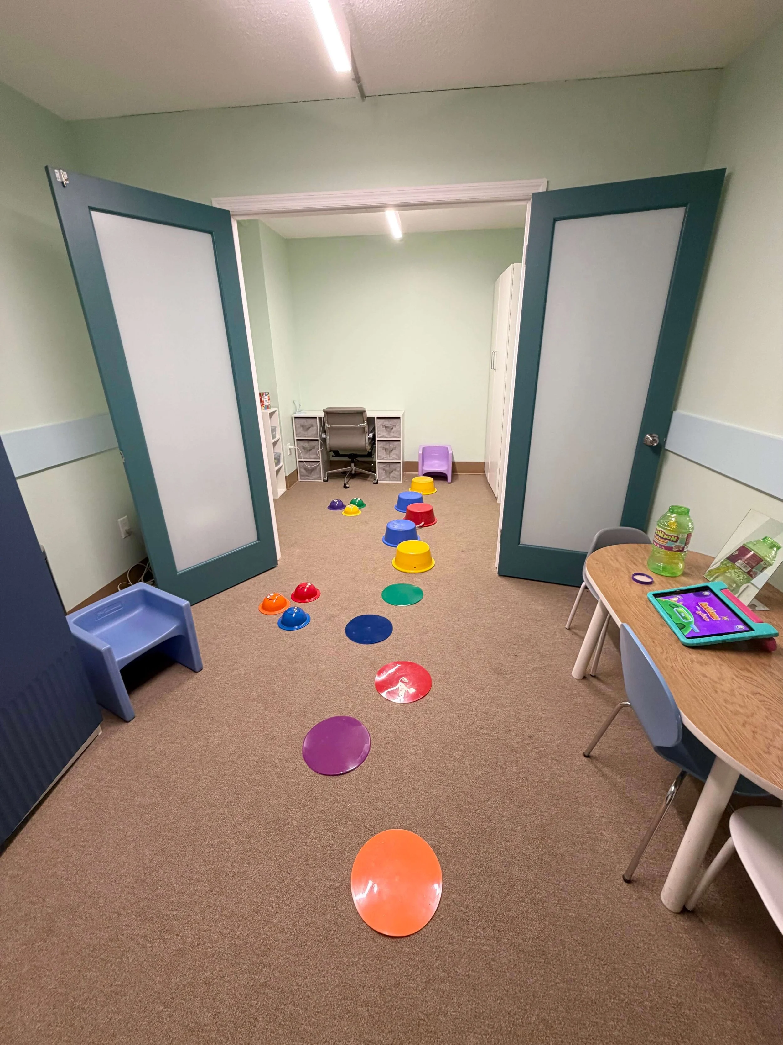 A classroom with colorful plastic bowls arranged in a line on the floor, a small table with children's items including a tablet, and a door opening into an office area with a desk and chair.