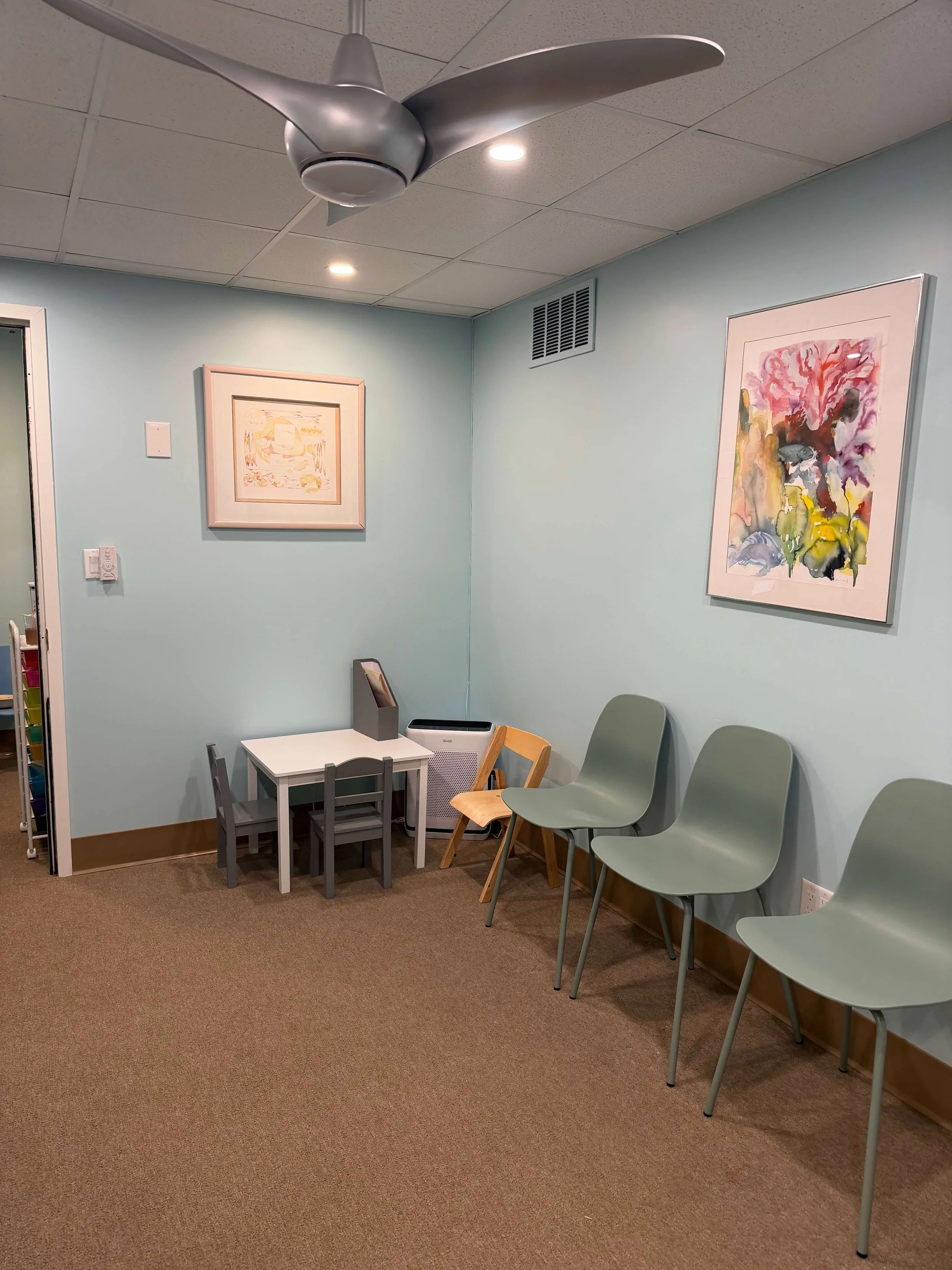 A waiting area with five light green chairs, a small white table with three gray chairs, framed artwork on light blue walls, and a ceiling fan.